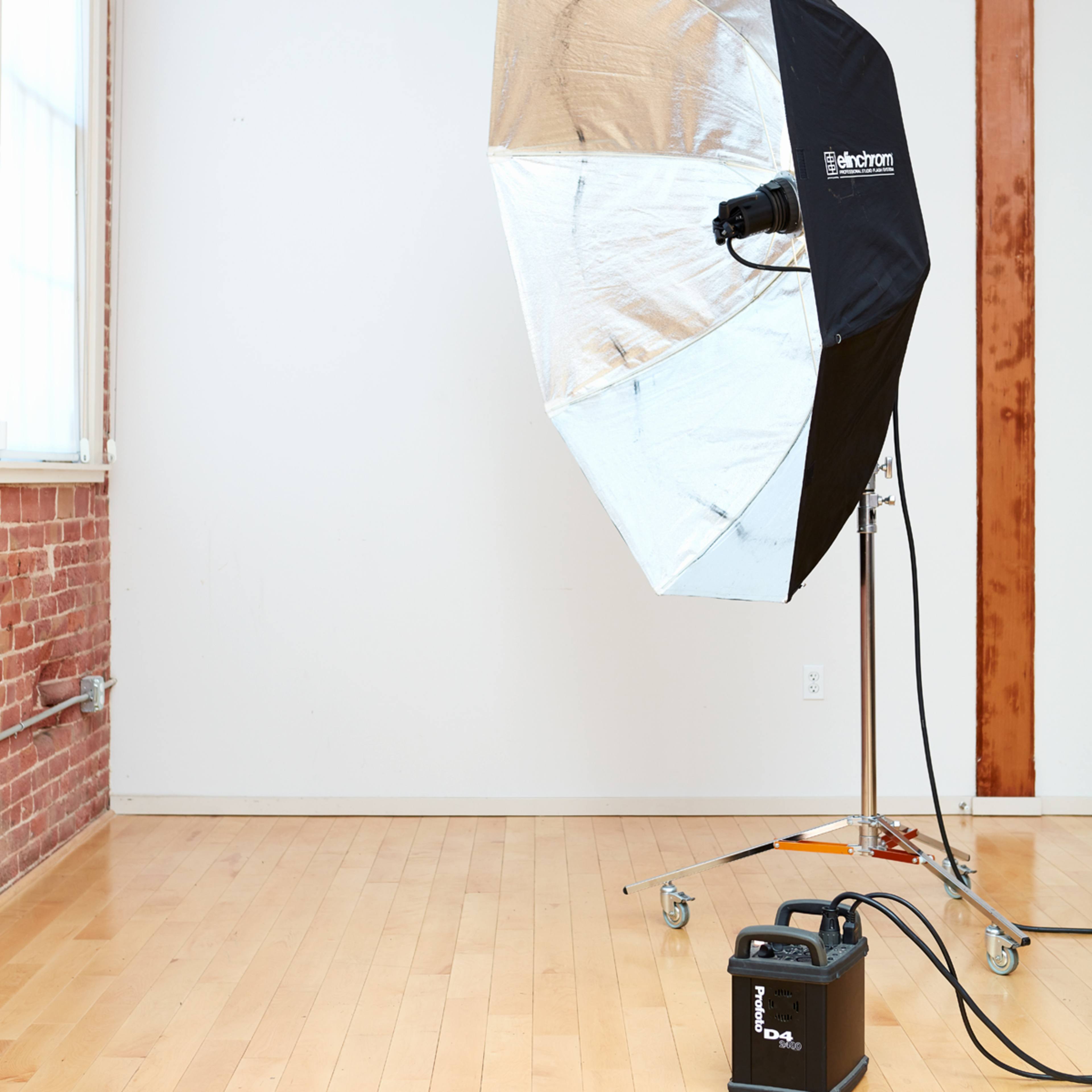 A large softbox light on wheels is positioned in a minimalist studio with wooden floors and exposed brick walls.