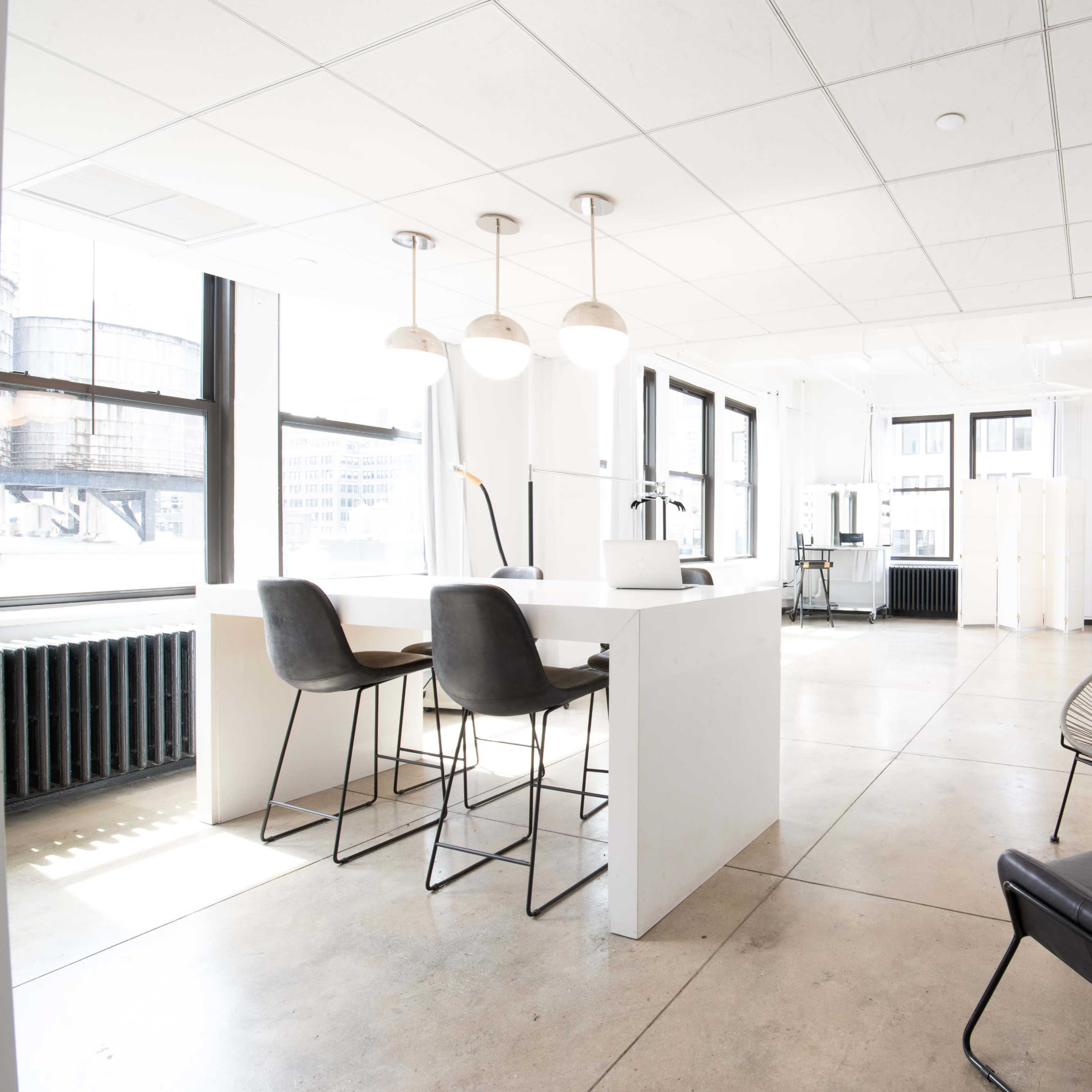 A modern, minimalist office space featuring a white countertop with bar stools, a black chair, and large windows allowing ample natural light.