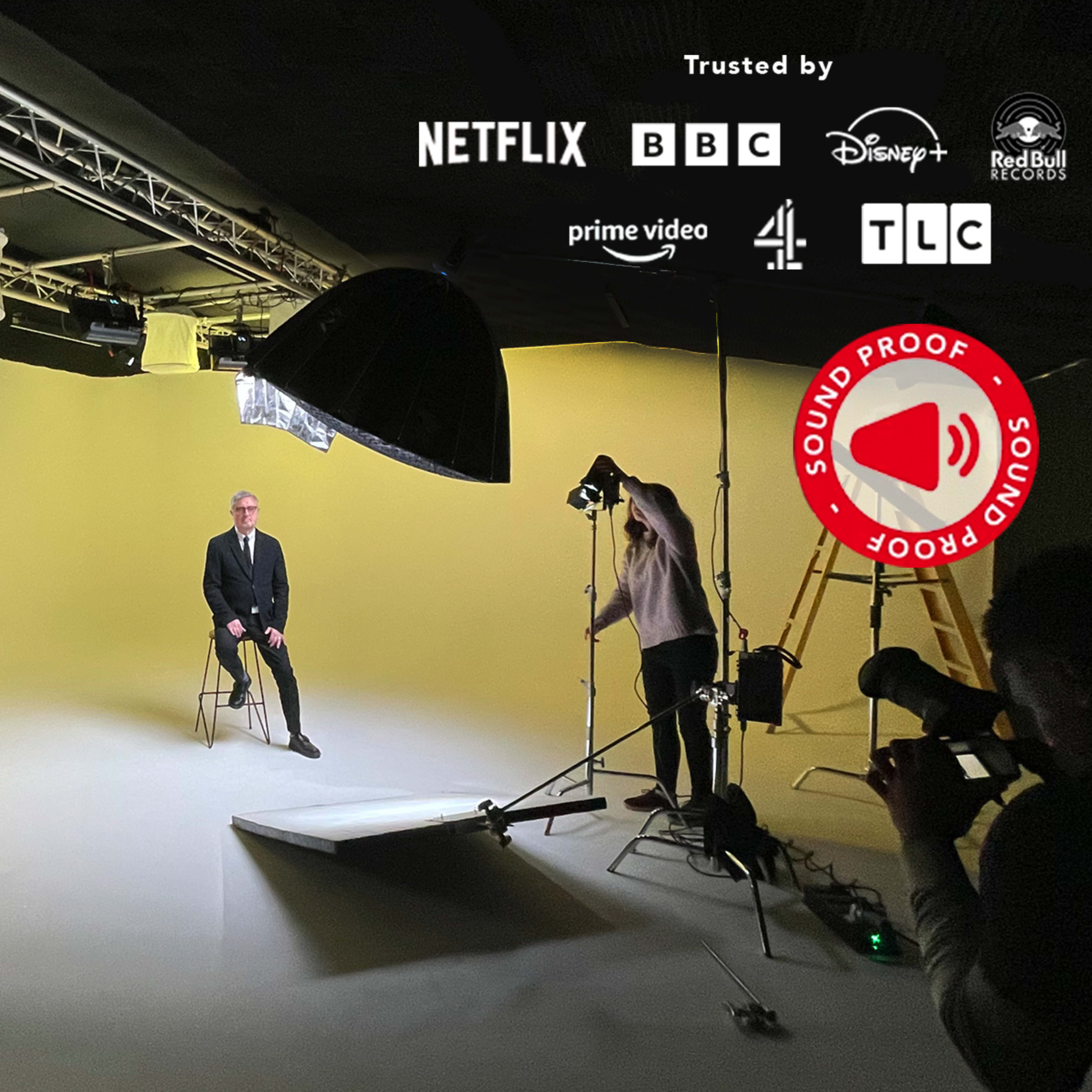 A studio setup features a man in a suit sitting on a chair in front of a yellow backdrop, with various lighting equipment and cameras positioned around him.