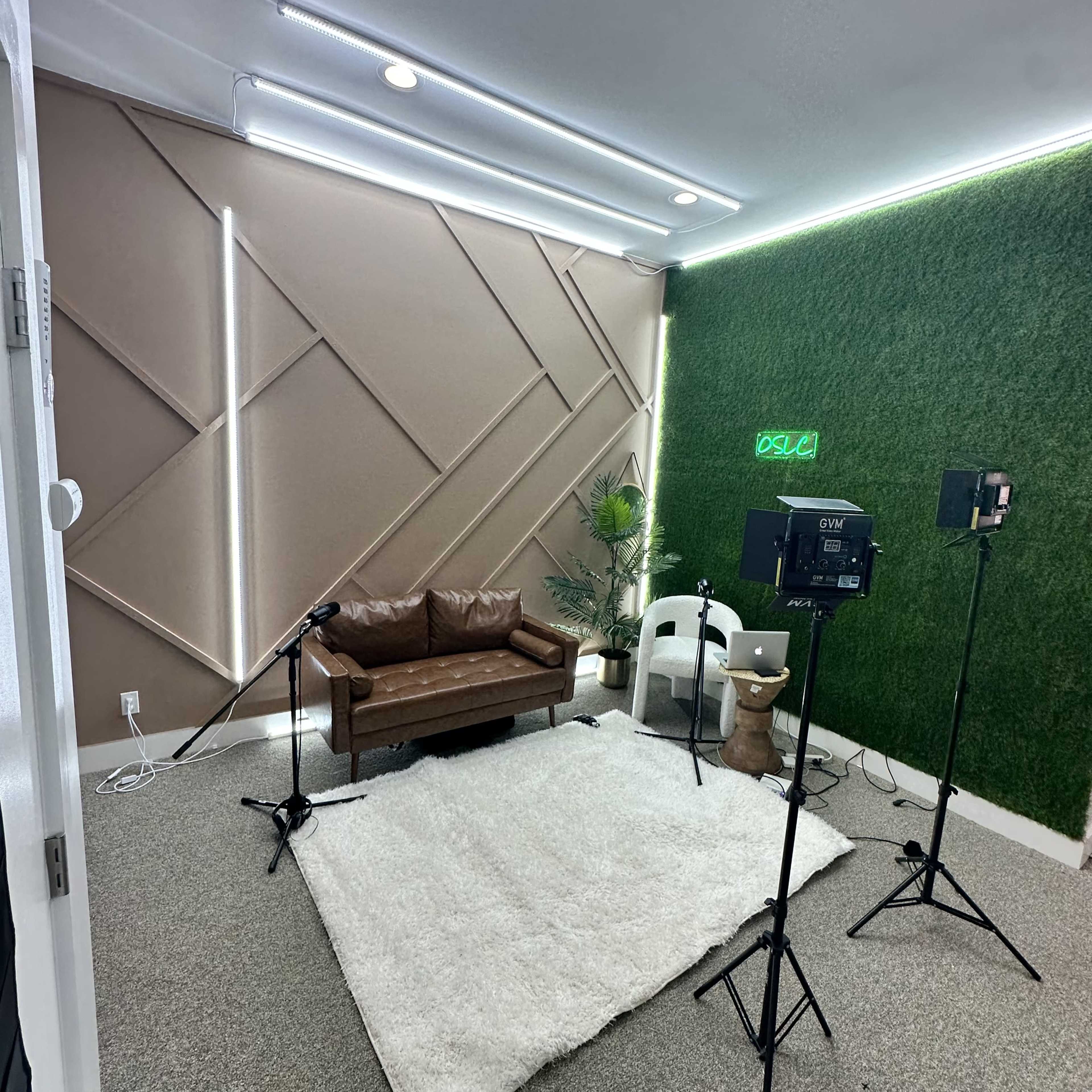 A modern recording studio featuring a brown leather sofa, a white rug, and lighting setup with cameras and equipment against a backdrop of green wall panels.