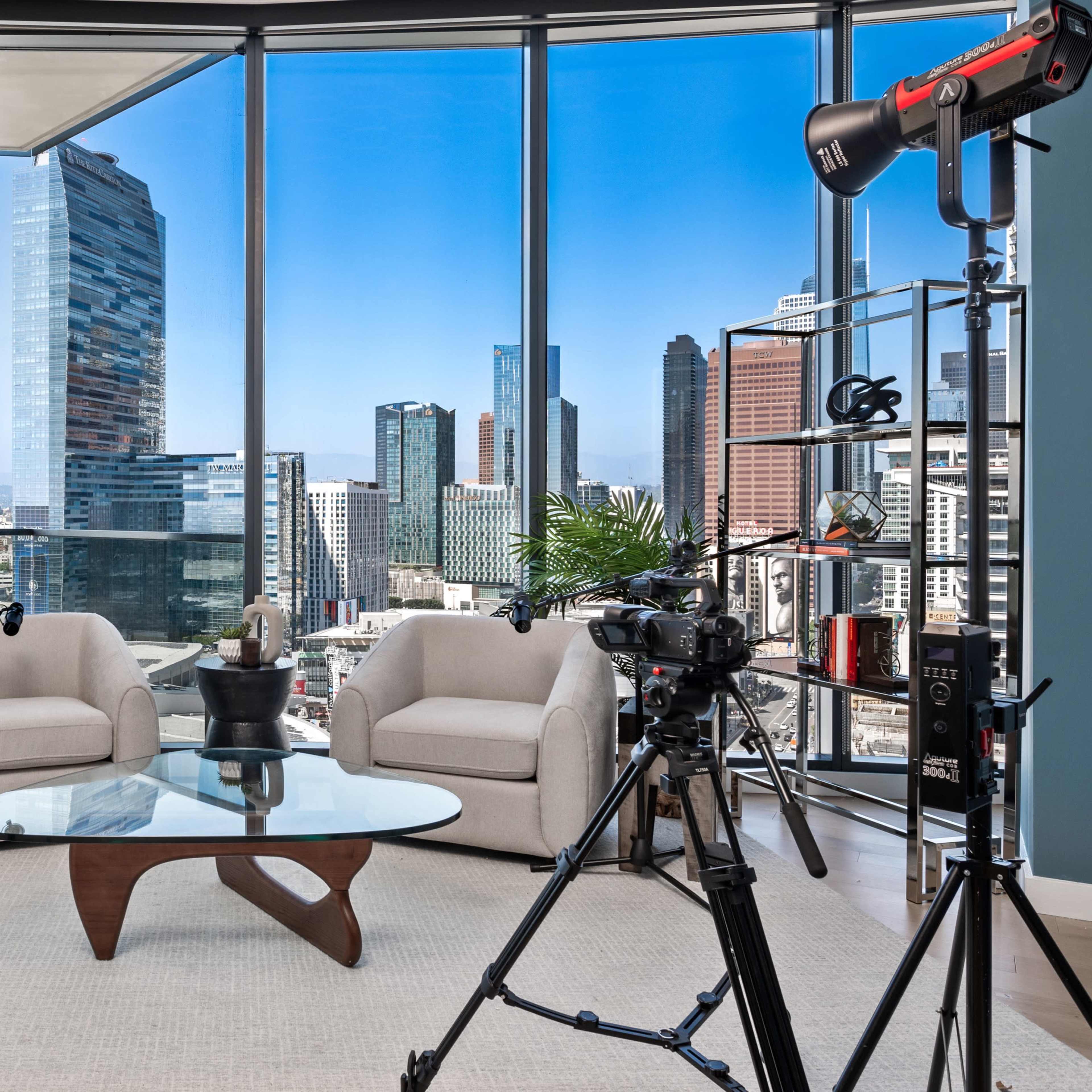 A modern studio setup with two chairs and a glass table, framed by large windows showing a city skyline and equipped with camera equipment and lighting.