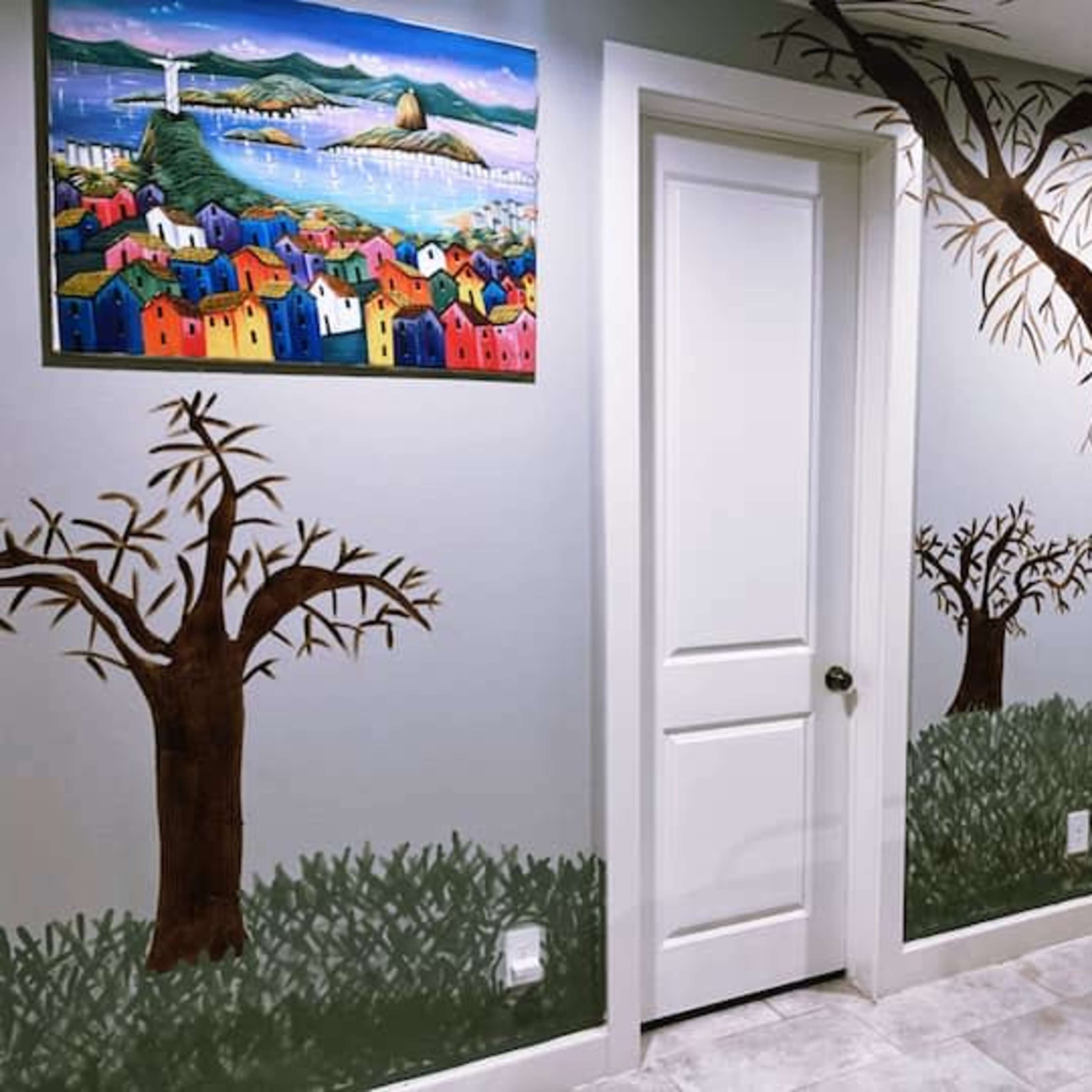A room with mural-painted trees and colorful houses, with a door and a large painting on the wall.