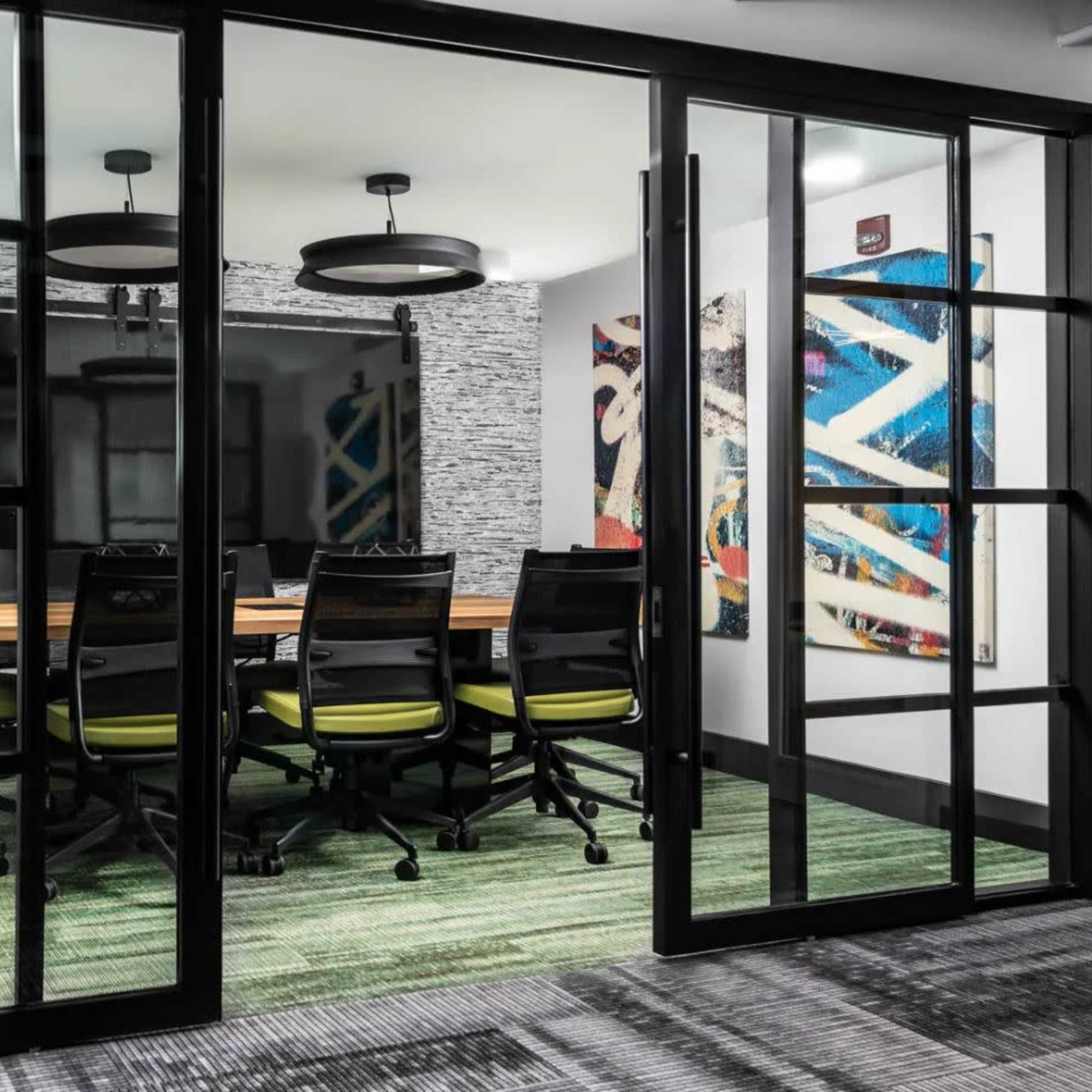 A glass-walled conference room features a wooden table surrounded by six black chairs with green accents, set against a backdrop of abstract artwork and a television screen.