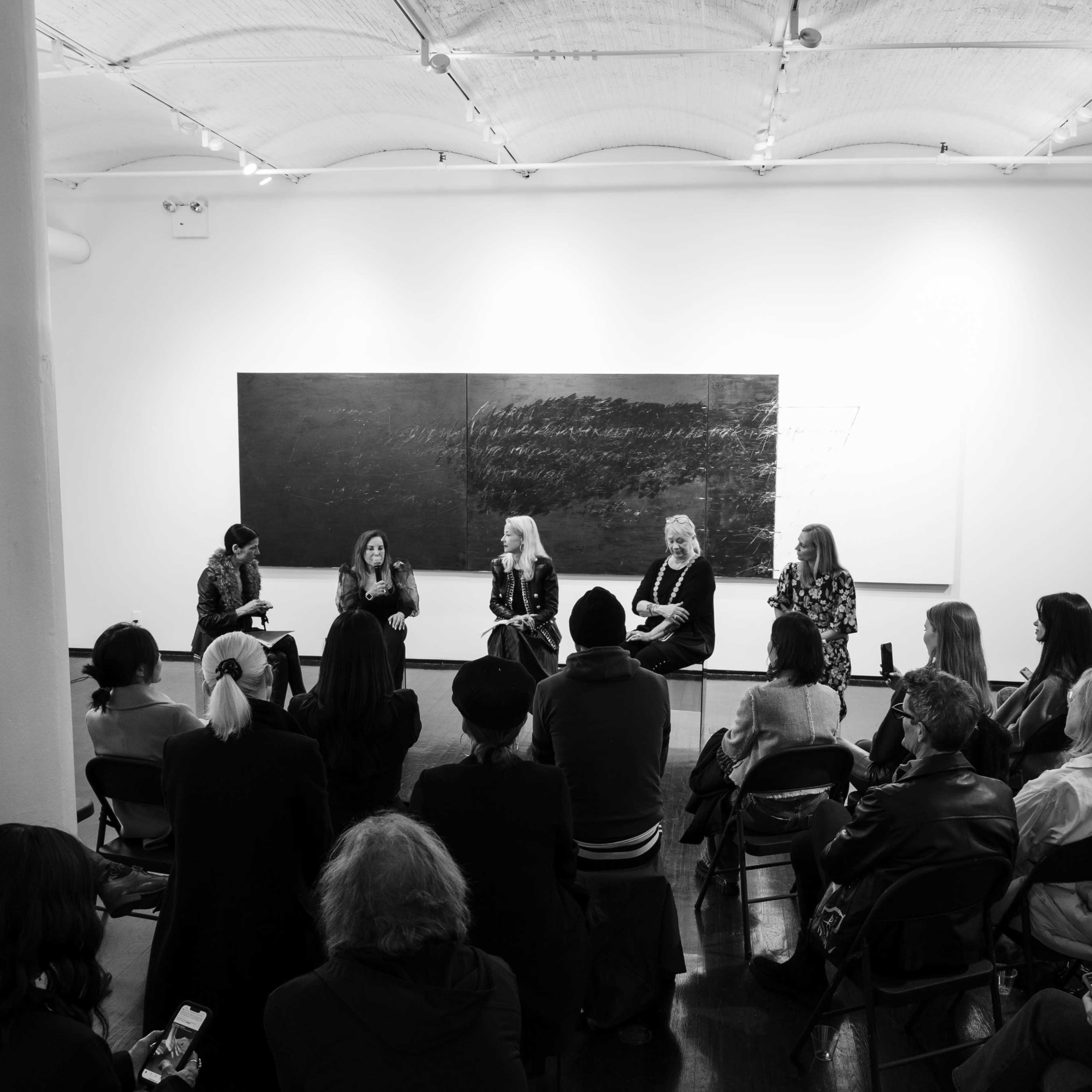 A panel discussion takes place in a gallery with four speakers seated in front of an audience.
