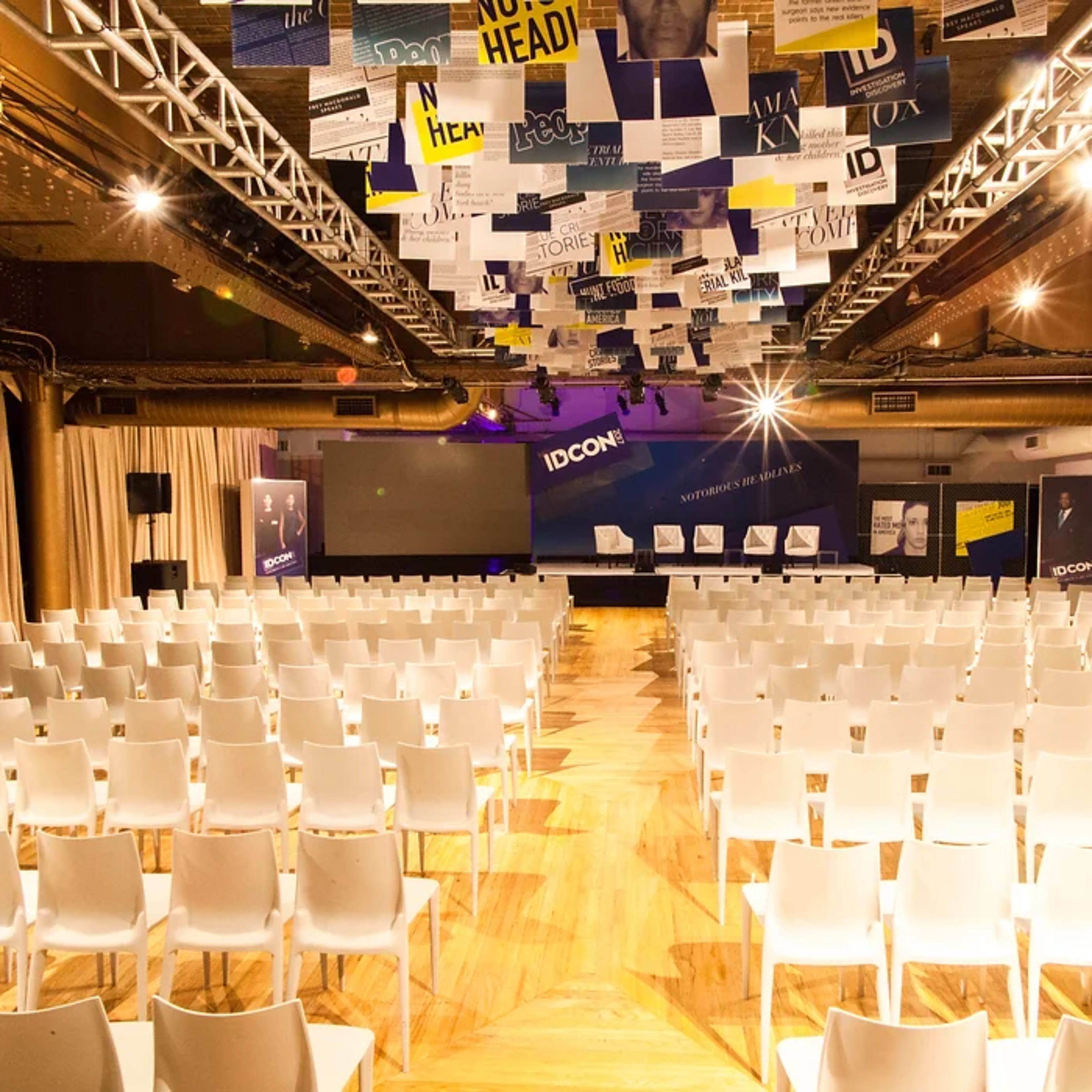 A conference hall is set up with rows of white chairs facing a stage adorned with banners and lights.