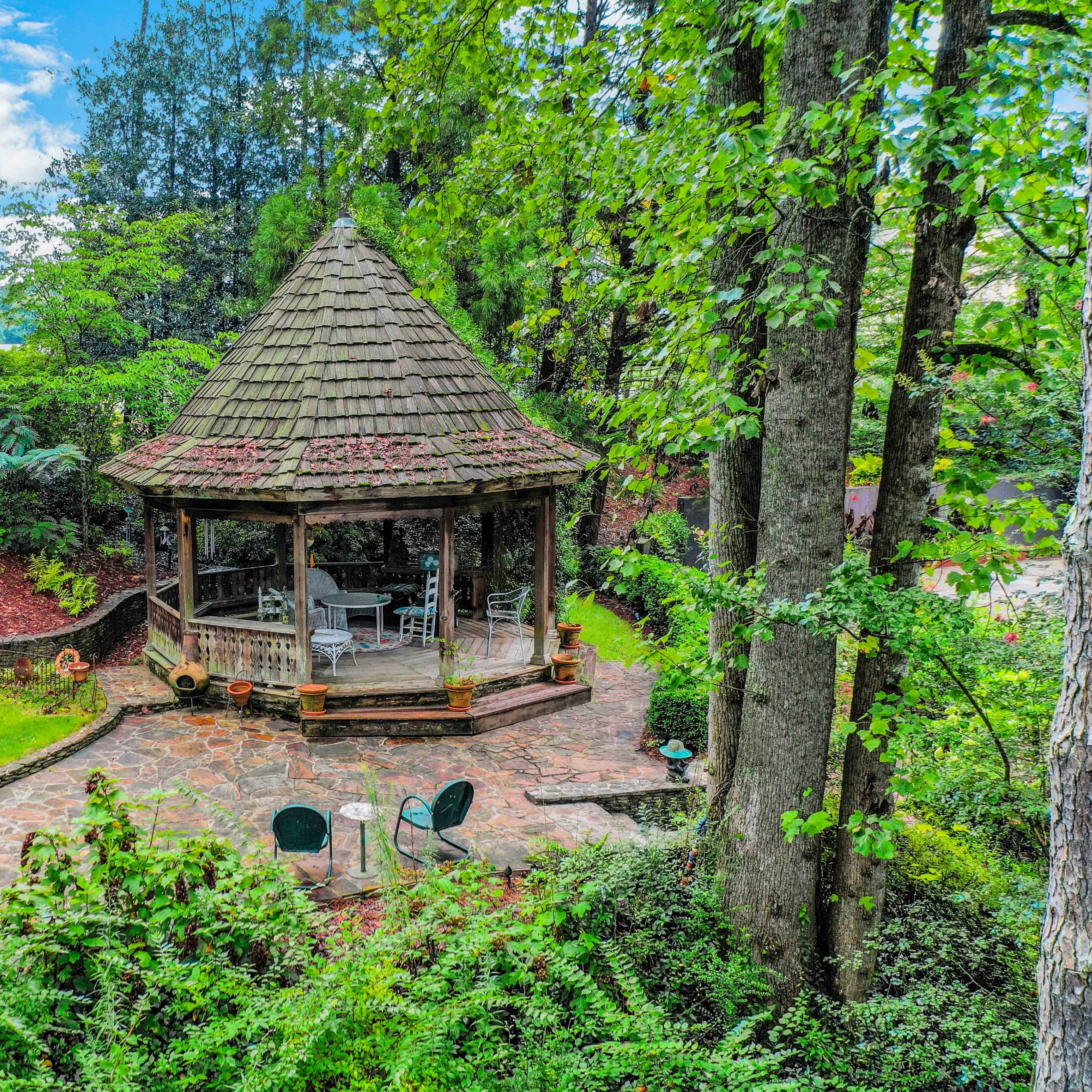A wooden gazebo with a shingled roof is surrounded by greenery and stone pathways in a landscaped garden.