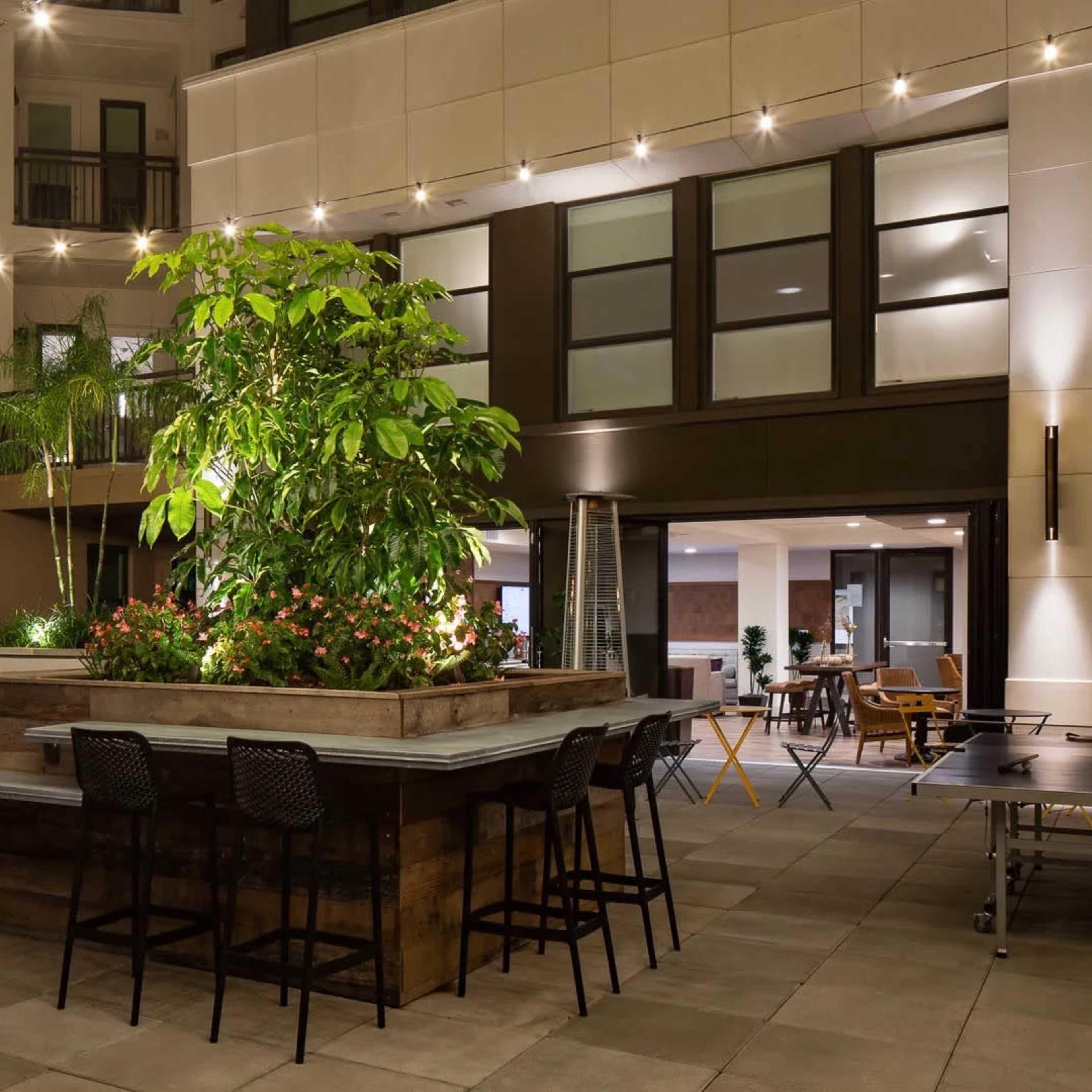 A patio area featuring a large potted plant arrangement, string lights overhead, and seating around a table under an umbrella.