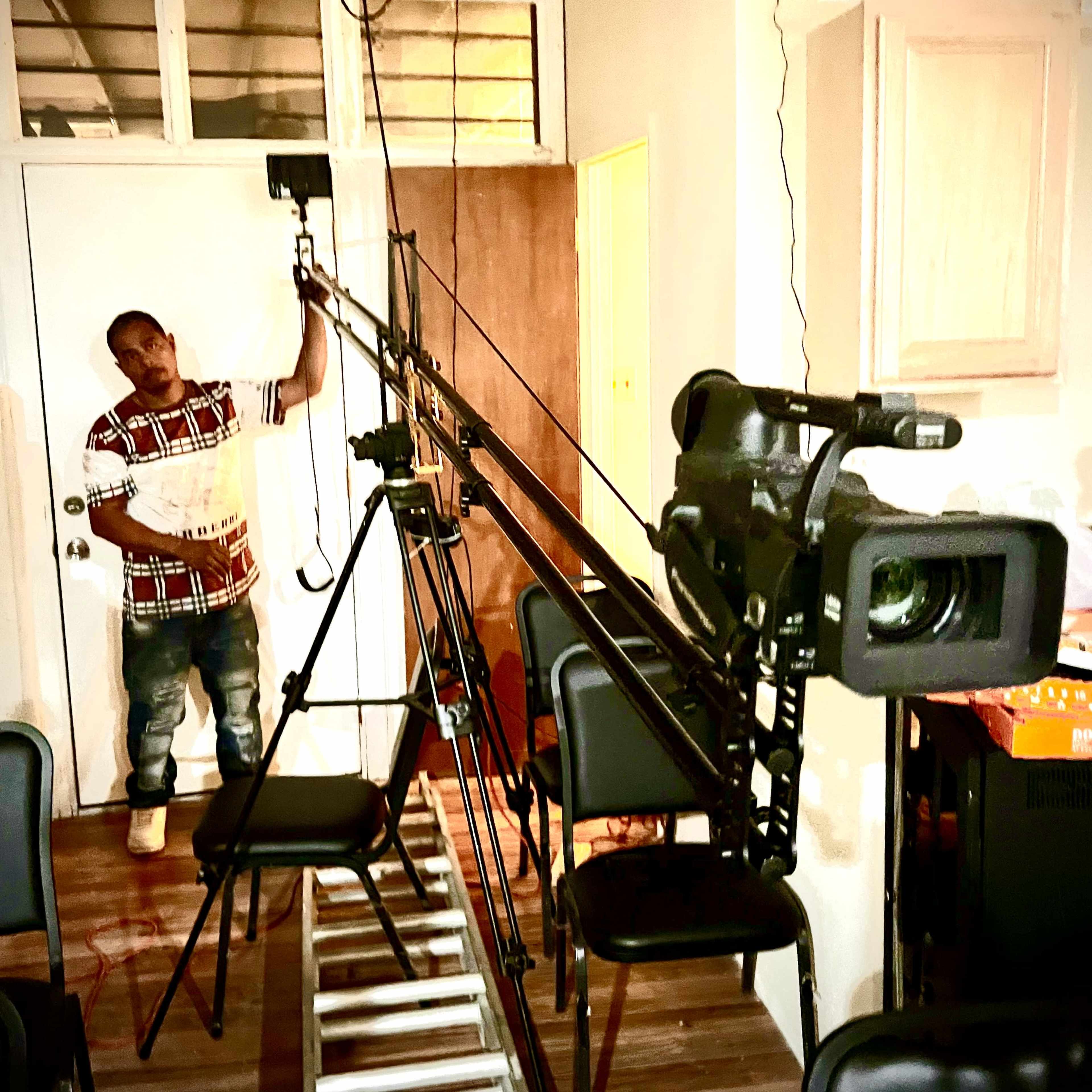 A man holds a clipboard while standing next to a camera setup with tripods and lighting in a room filled with chairs.