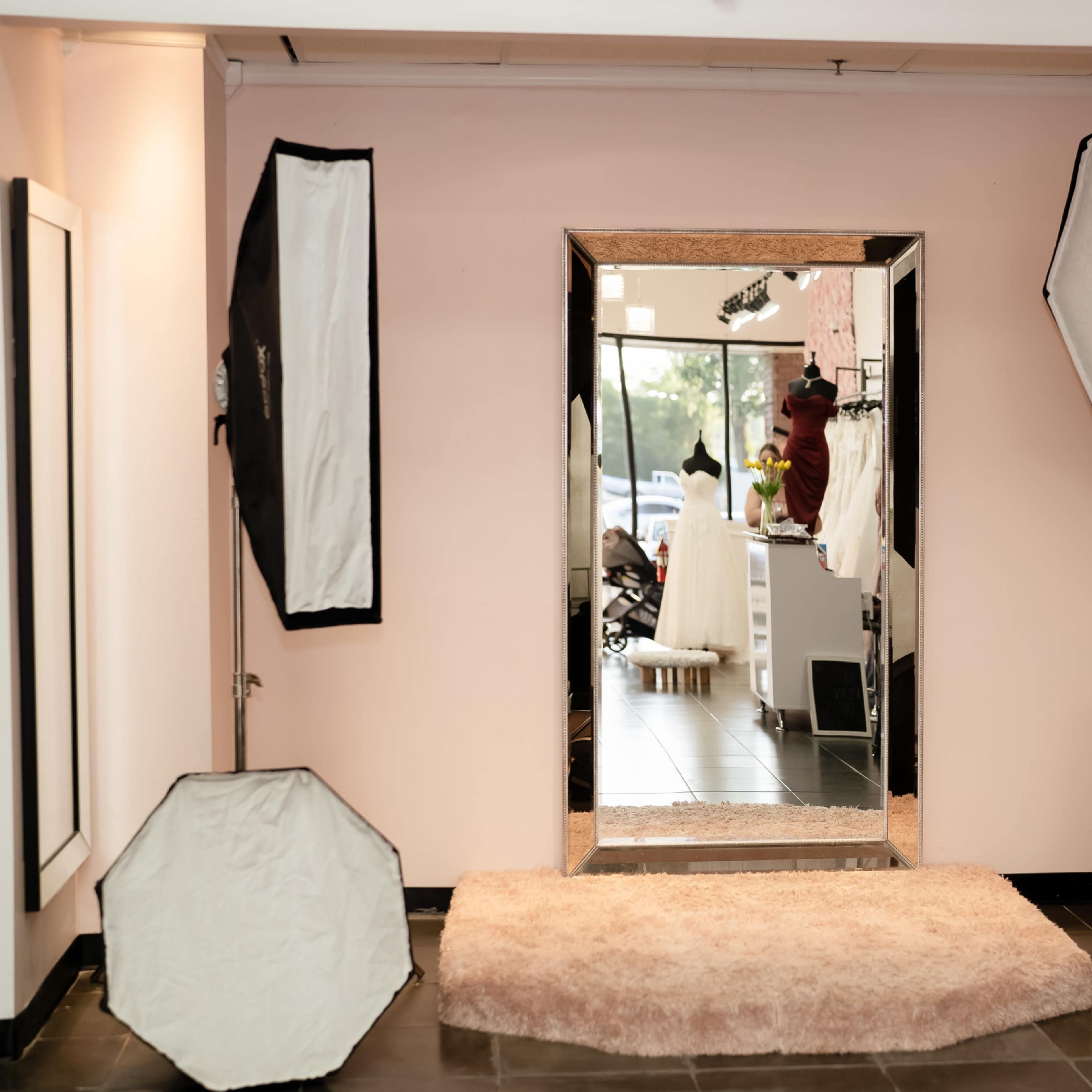 A bright, modern fitting room with a large mirror surrounded by soft light reflectors on either side, set against pink walls and a black tiled floor.