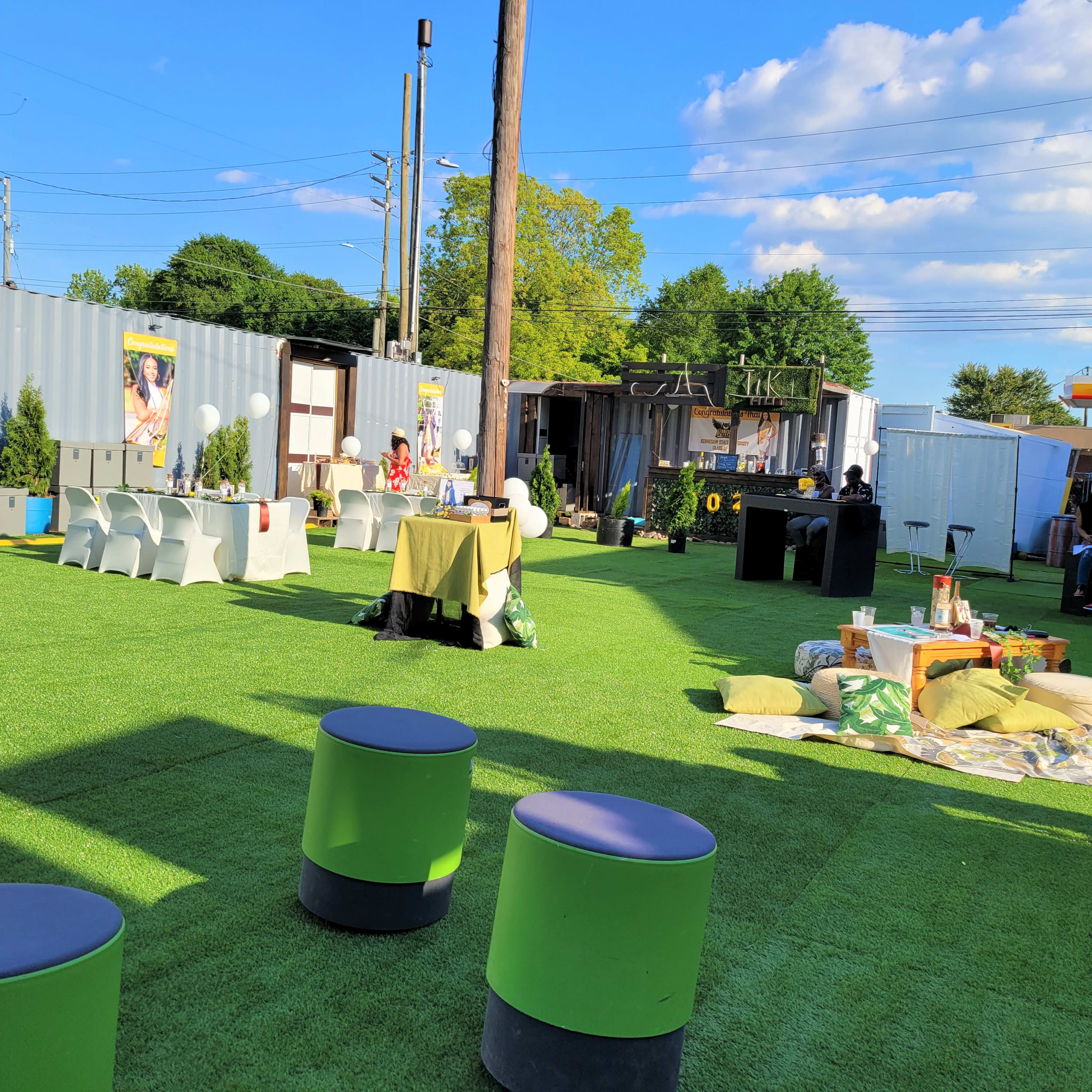 An outdoor event space with green artificial turf, seating arrangements, and shipping containers in the background.