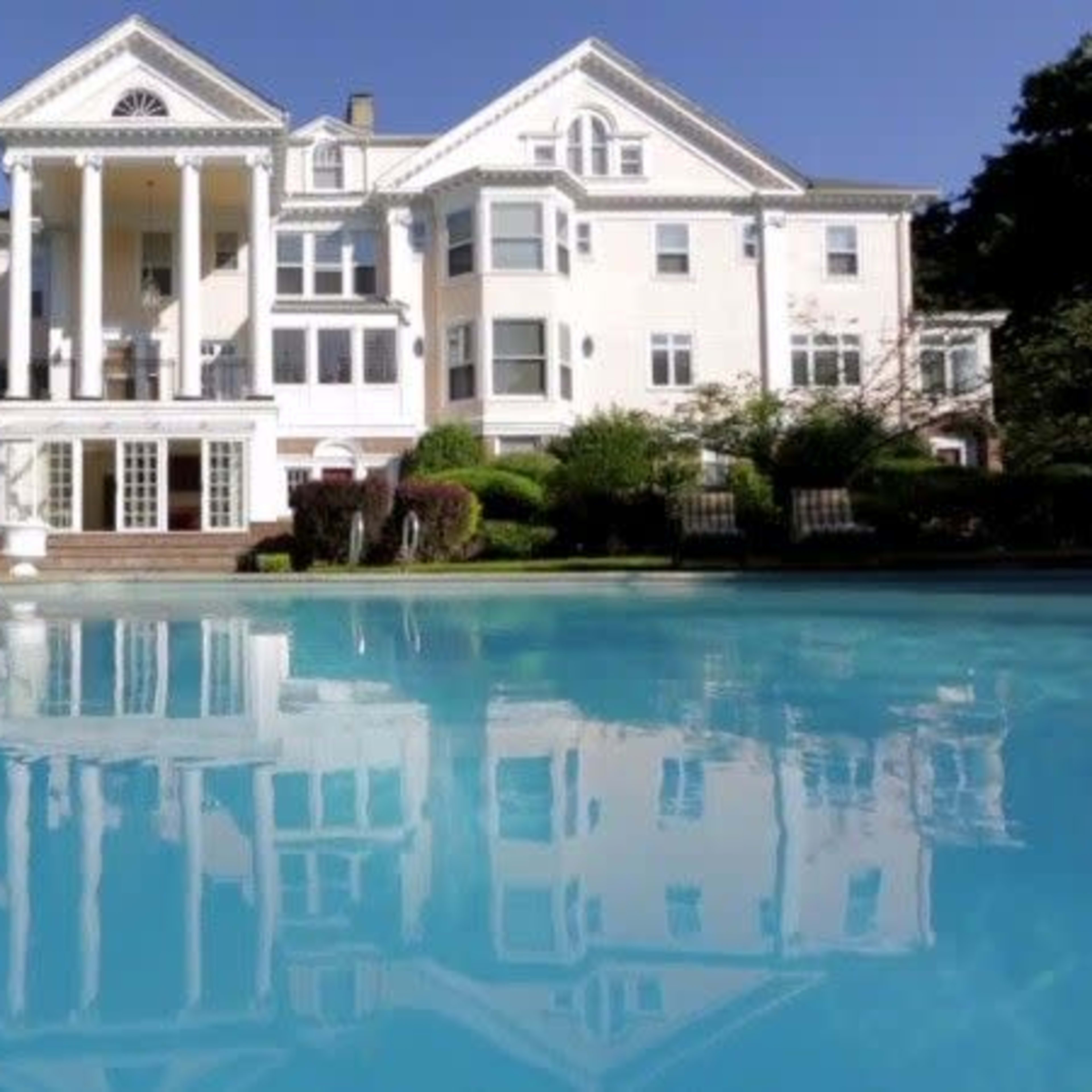 A large, white mansion with columns is reflected in a clear blue swimming pool surrounded by greenery.