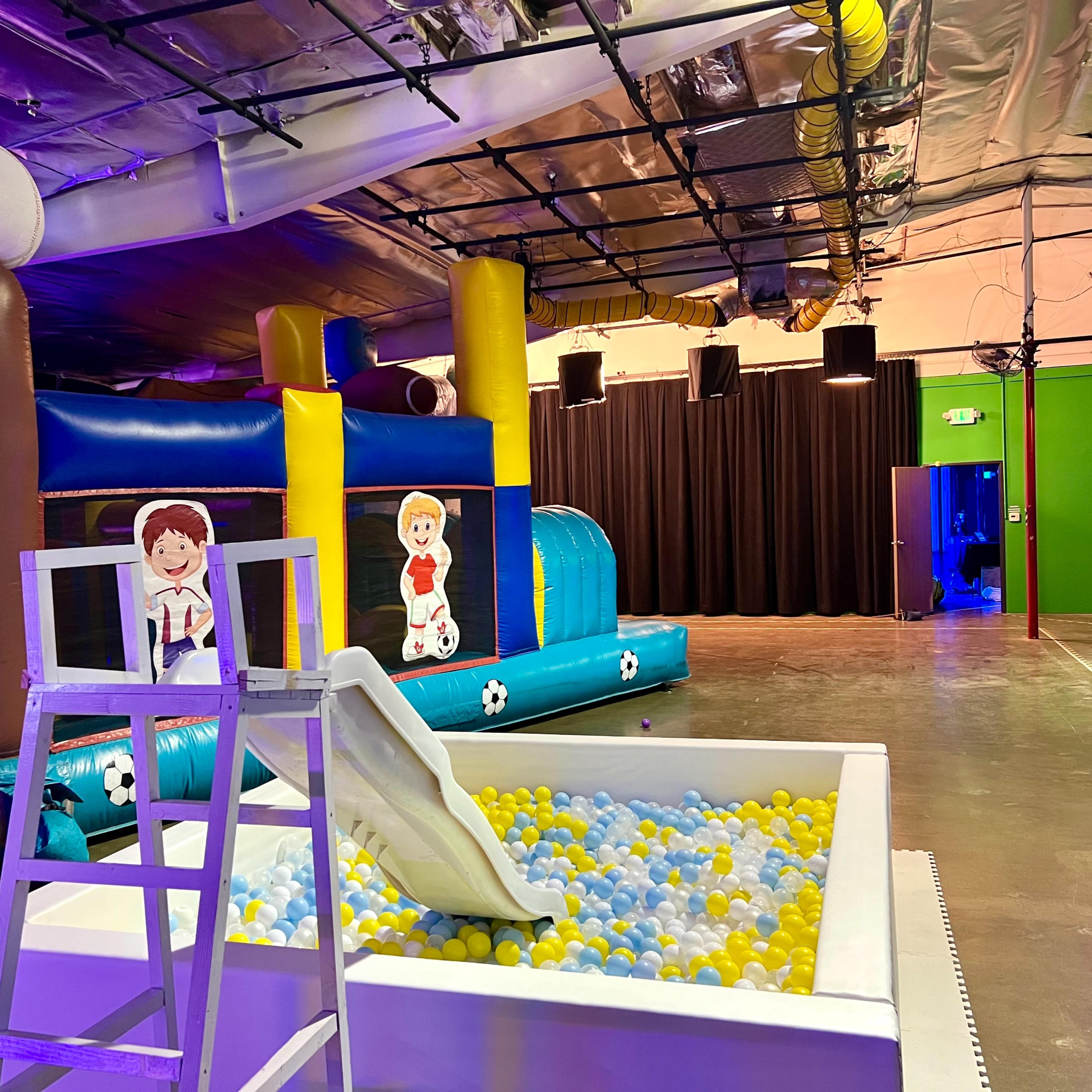 A colorful indoor play area featuring inflatable bounce structures, a slide, and a pool filled with blue and yellow balls.
