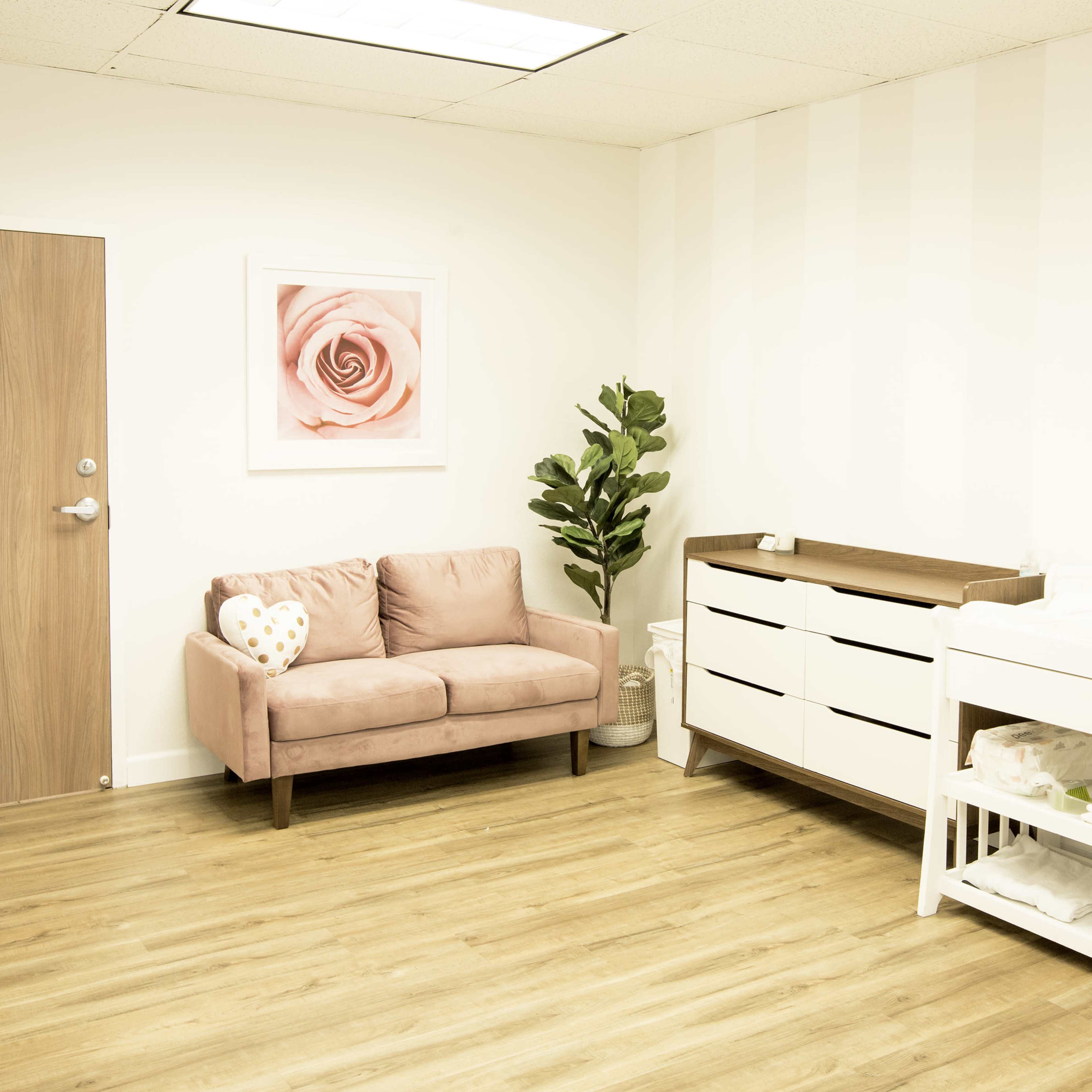 A bright room with wooden flooring, featuring a pink couch, a dresser, a changing table, and a leafy indoor plant against a striped wall.