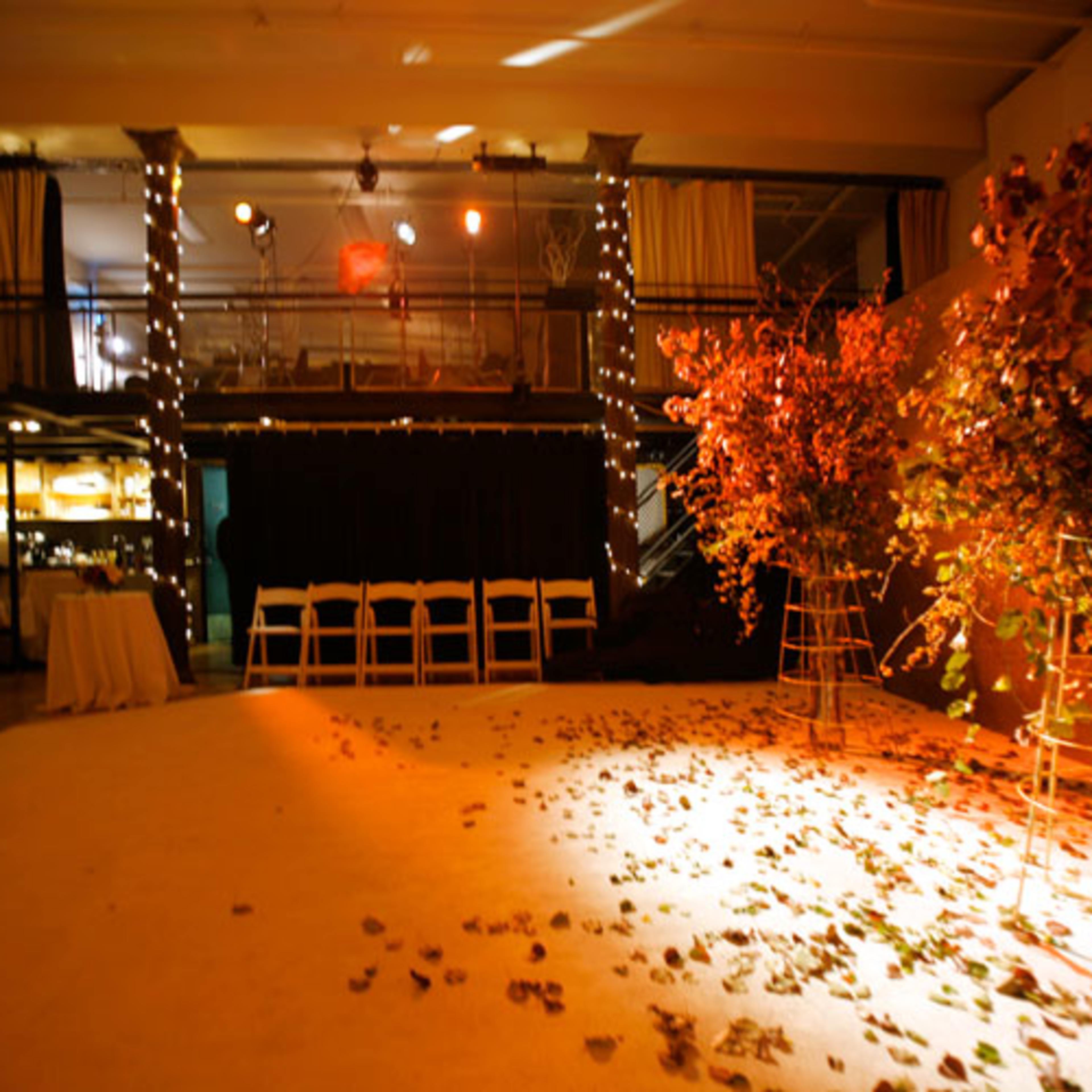 The scene shows an event space decorated with string lights, two large floral arrangements on stands, and scattered leaves on the floor.