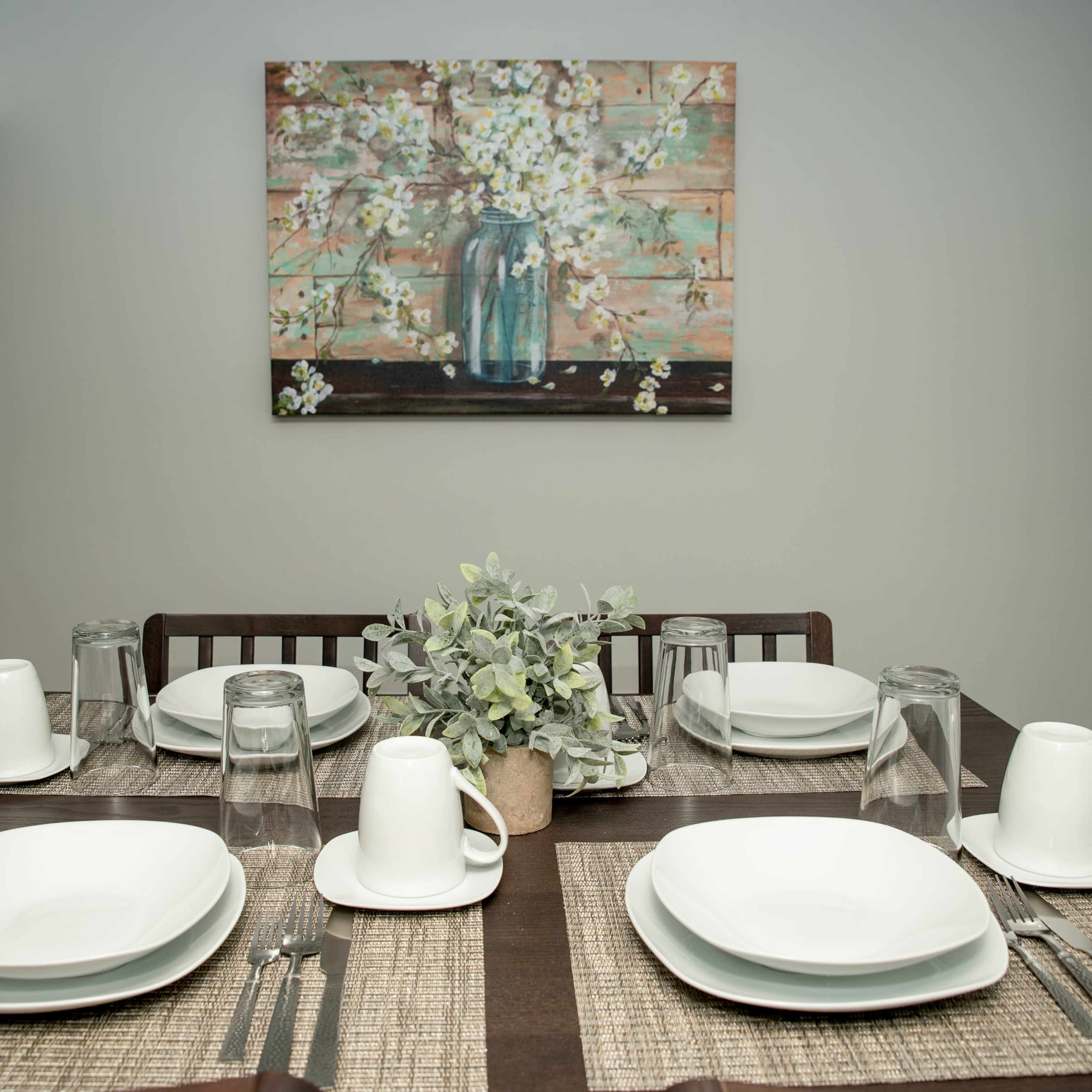 A dining table is set with white dishes, glasses, and a floral centerpiece against a wall featuring a painting of flowers in a jar.