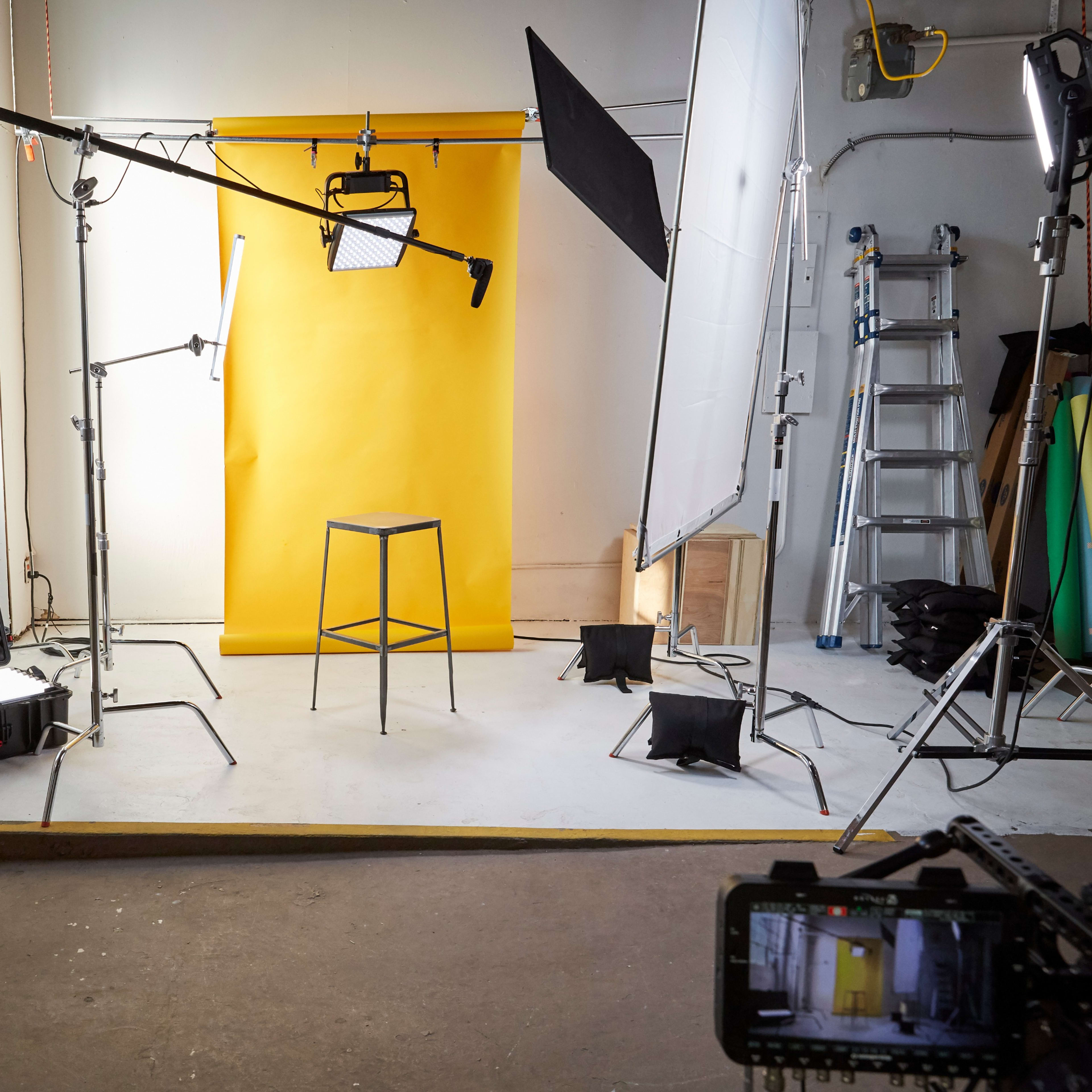 A photography studio is set up with a yellow backdrop, a black stool, and various lighting equipment arranged around the space.