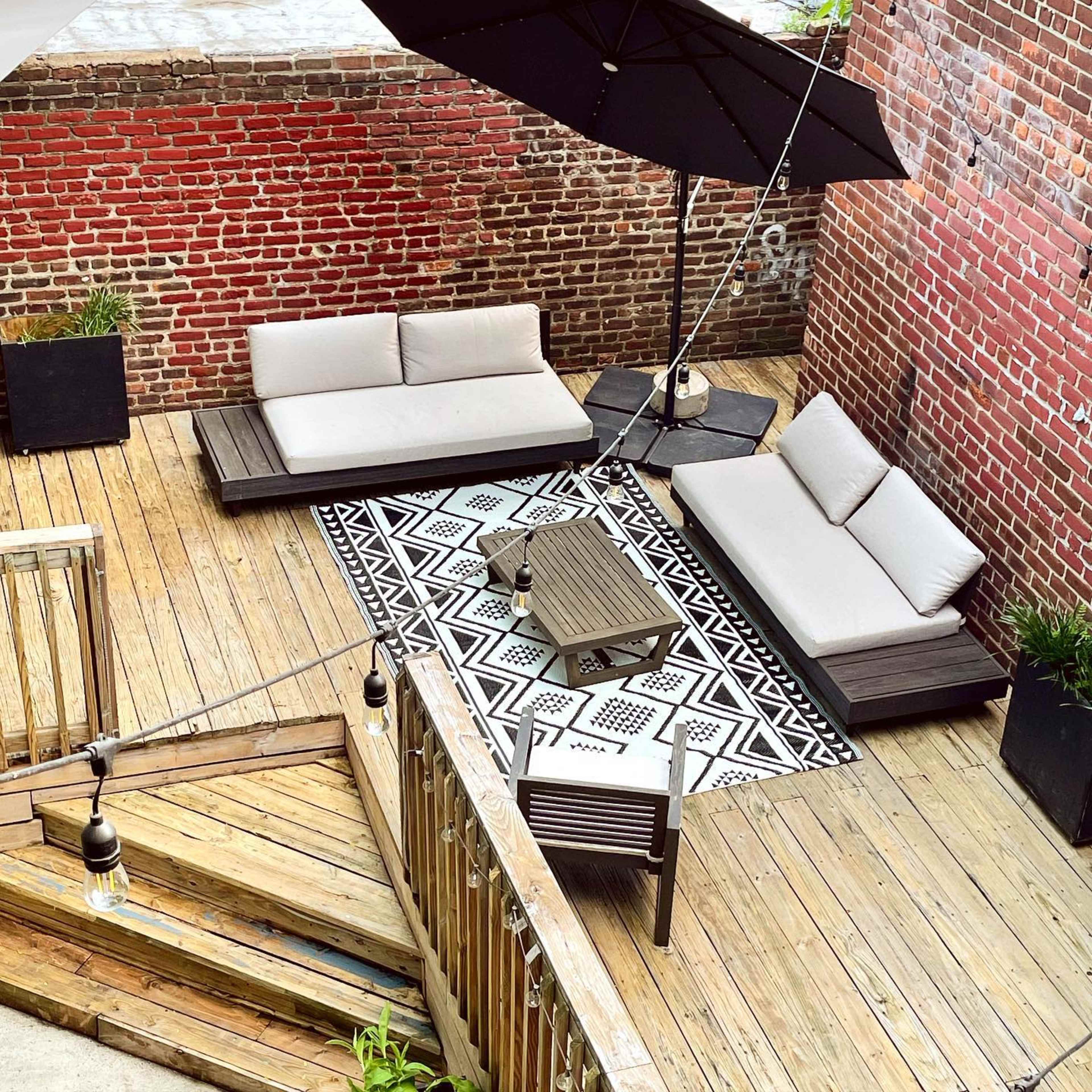 A wooden deck area furnished with outdoor couches, a patterned rug, a coffee table, and a large umbrella, surrounded by brick walls and potted plants.