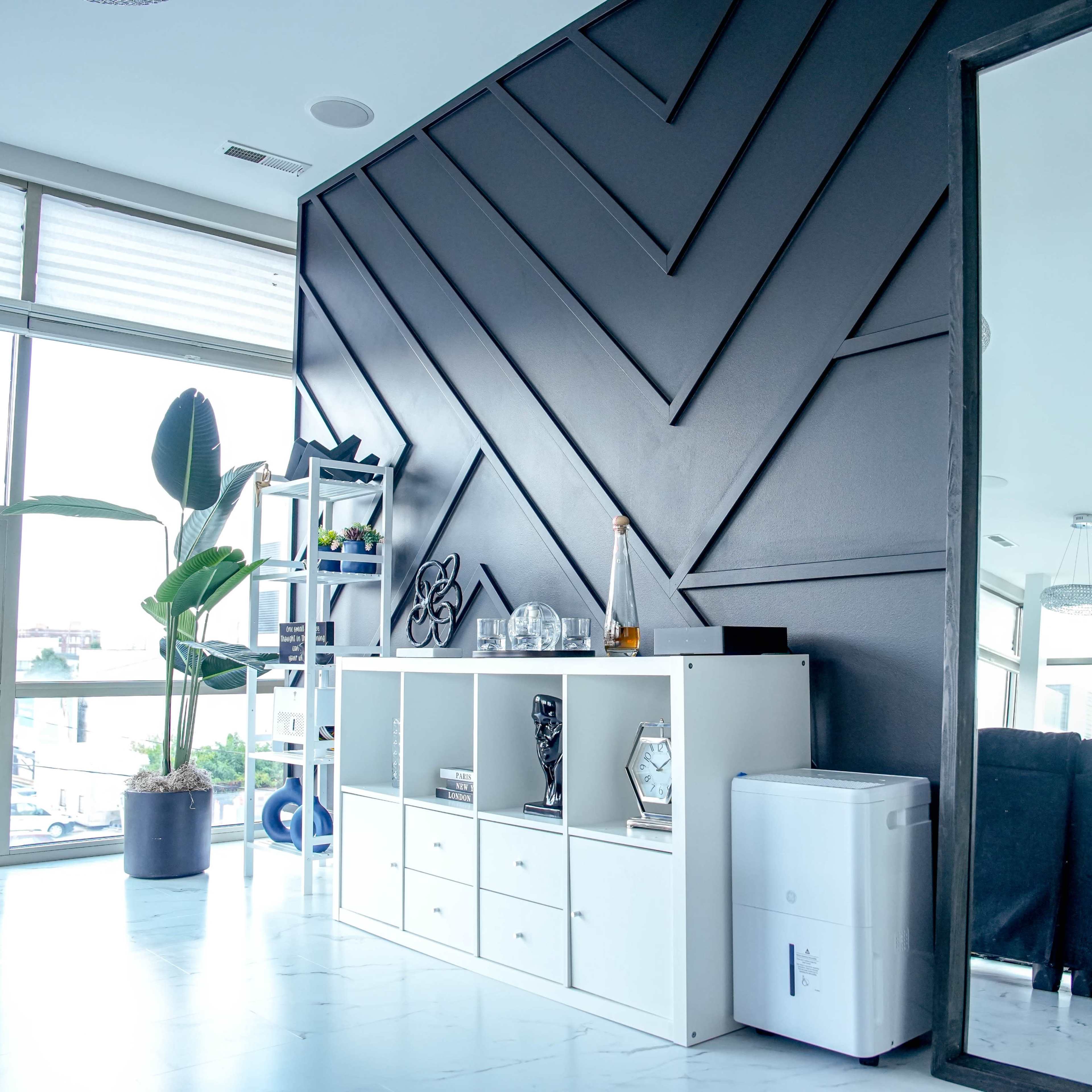 A modern interior room with a black accent wall, a white cabinet displaying decorative items, a potted plant, and a large mirror.