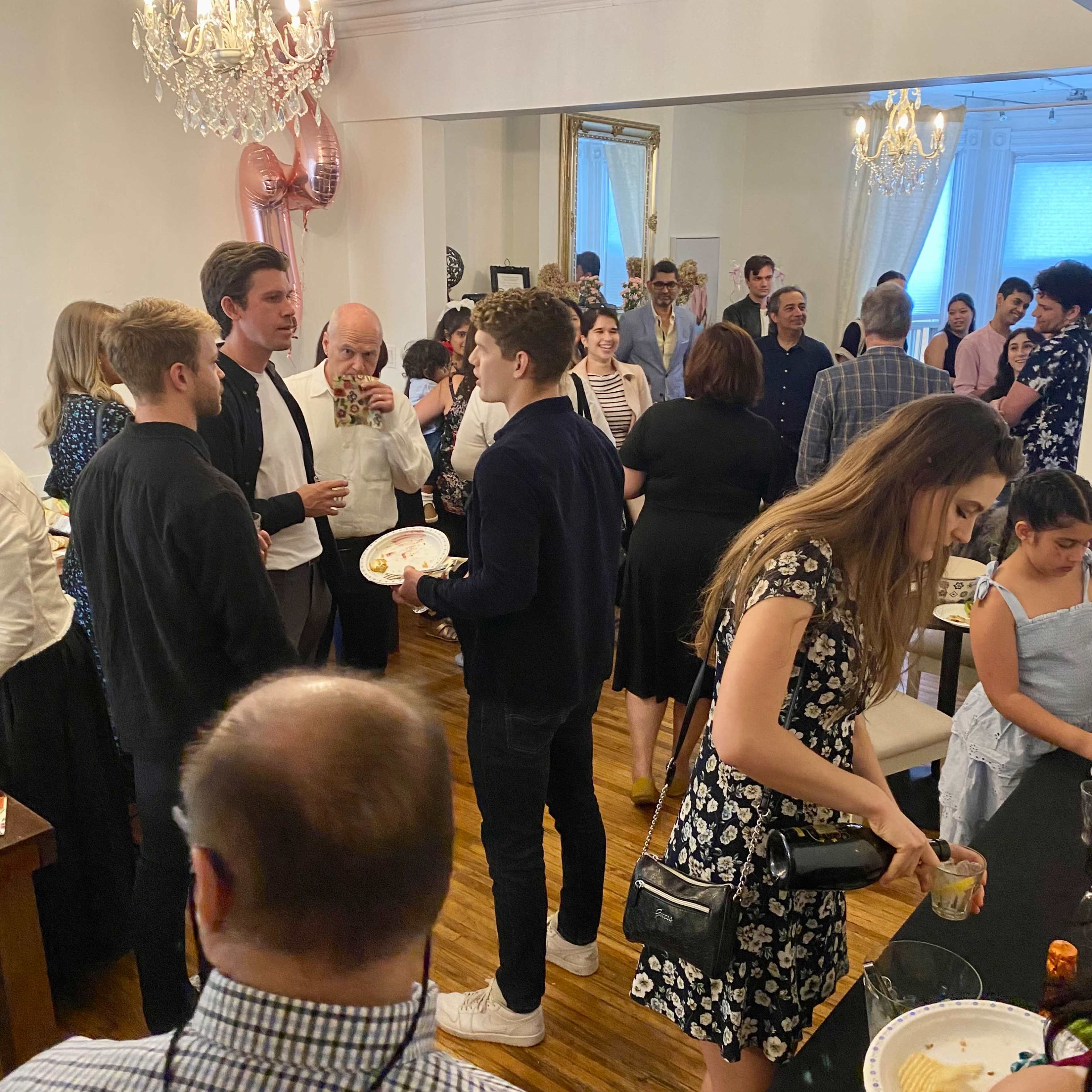 A crowd of people mingles at a social gathering in a well-lit room adorned with a chandelier and decorations.