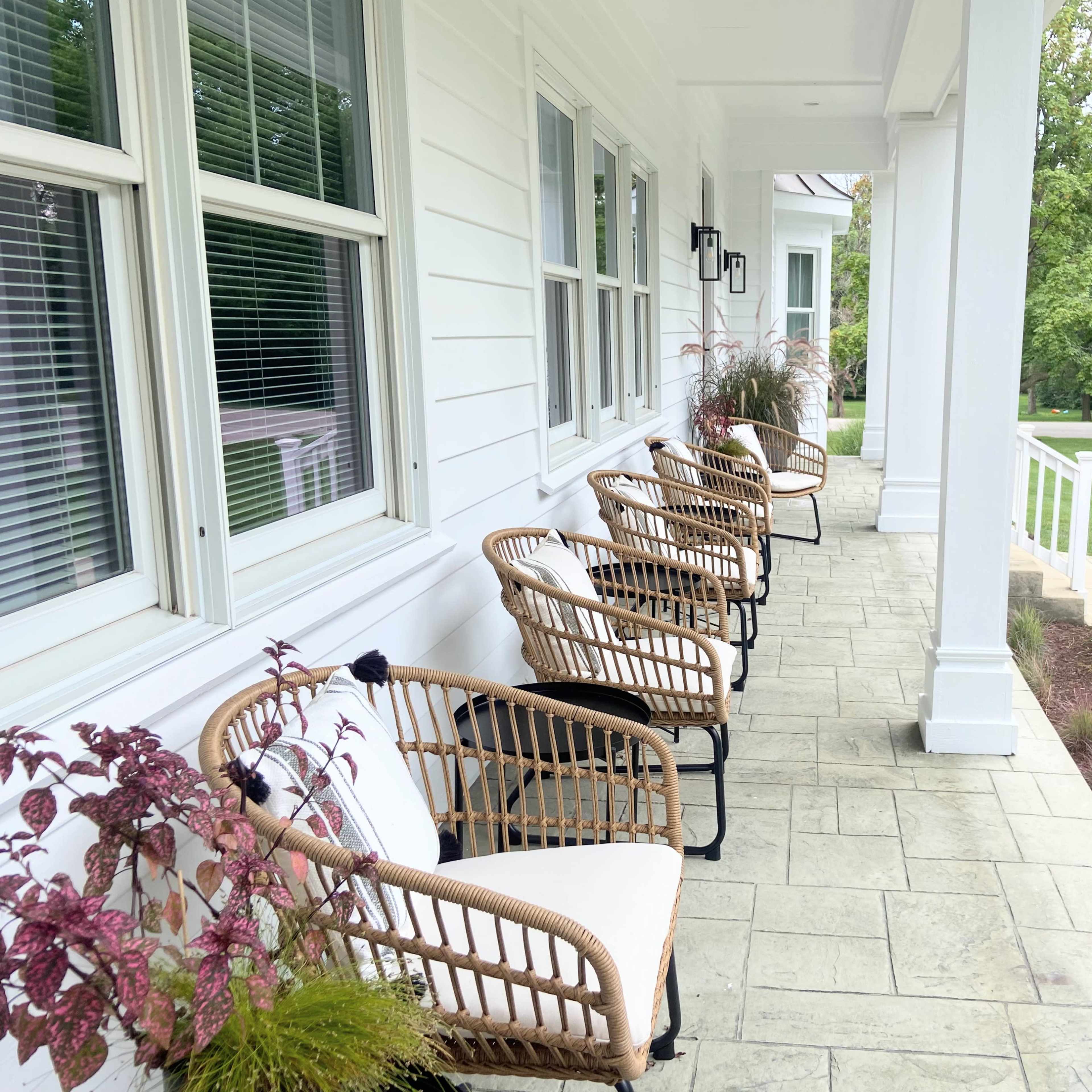 A row of wicker chairs is positioned on a covered porch beside potted plants and a grassy lawn.