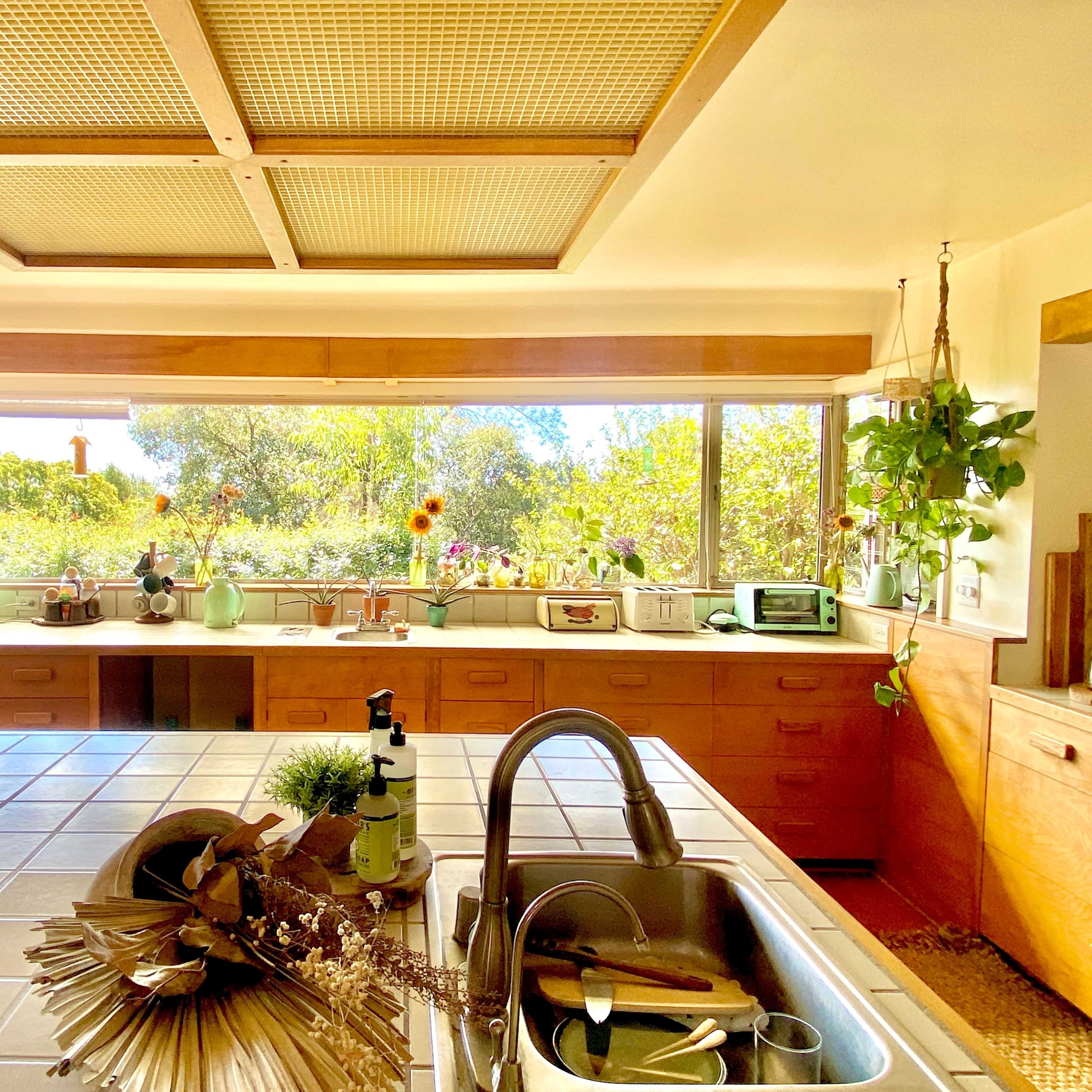 A bright kitchen features a tiled countertop, wooden cabinetry, and large windows showcasing greenery outside.