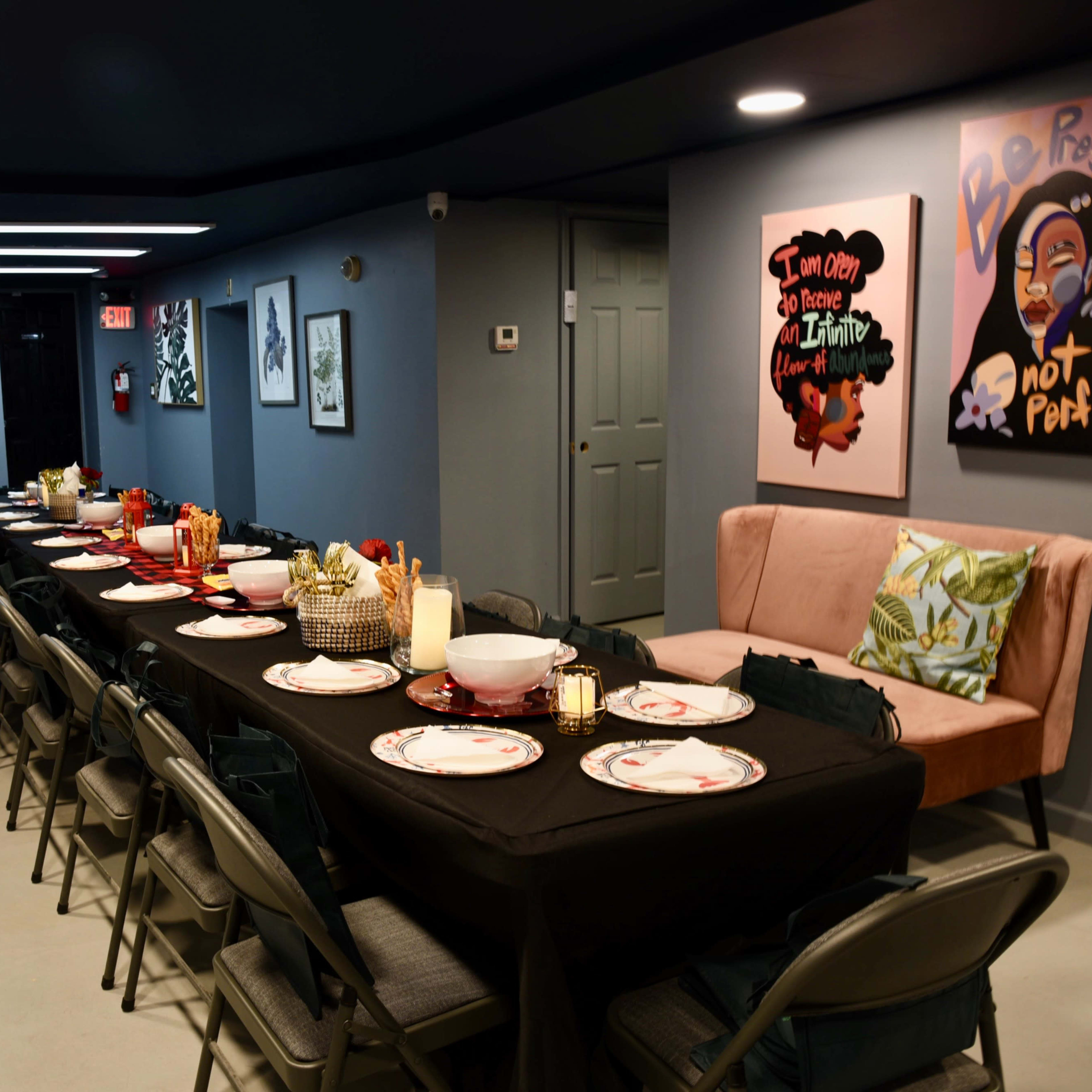 A long table is set for a meal, decorated with plates and glasses, and surrounded by folding chairs in a dimly lit room with artwork on the walls.