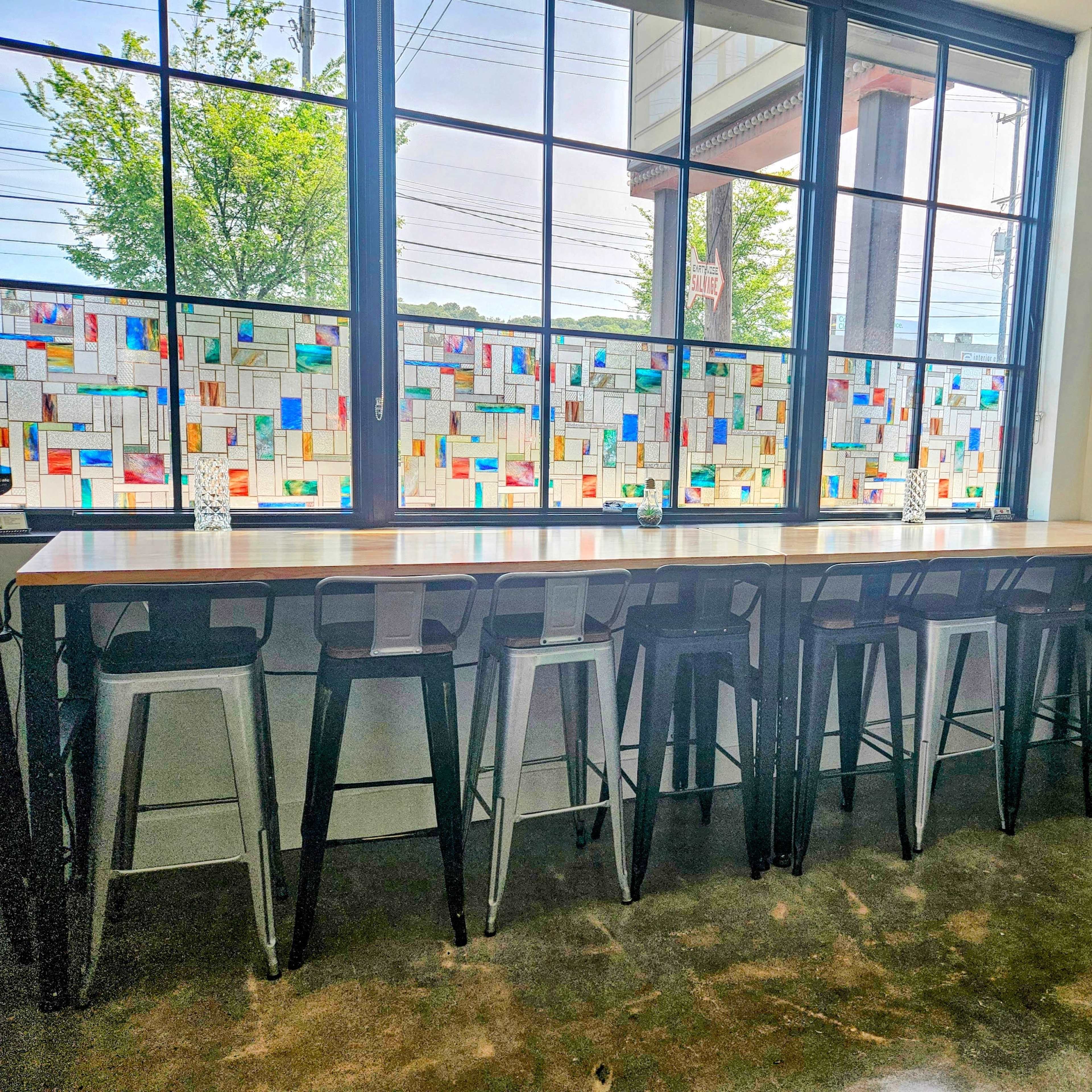 A modern cafe interior with a long wooden counter and metal stools, backed by colorful stained glass windows and a brick wall.