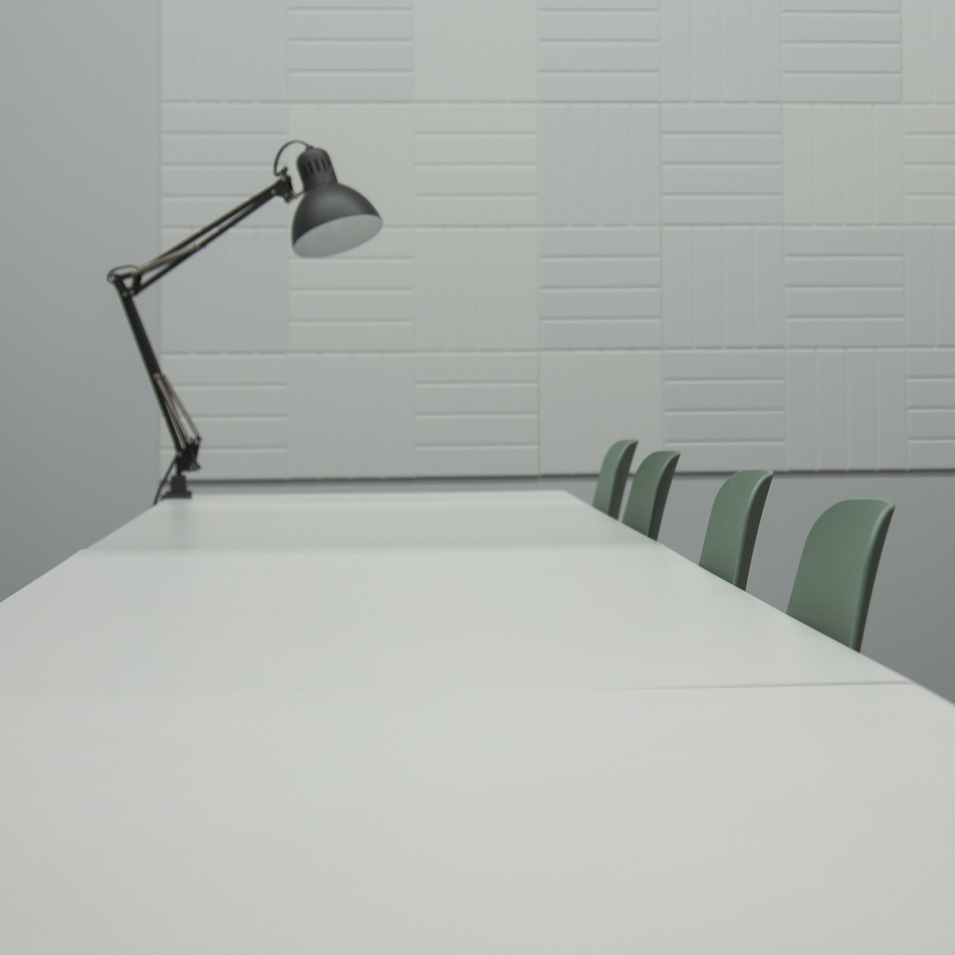 A row of green chairs sits at a long, white table in a minimalist room with a desk lamp and textured wall.