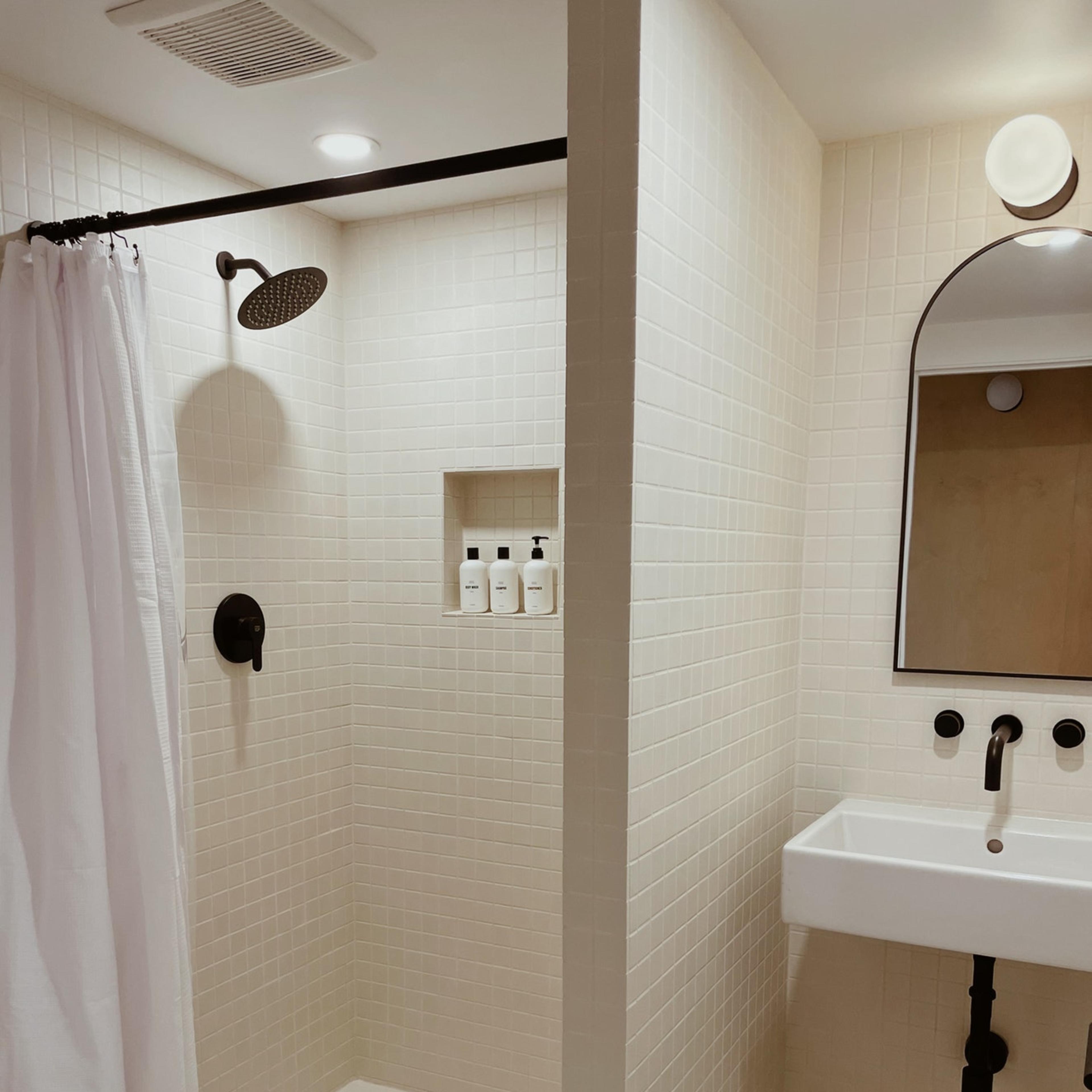 A modern bathroom with a tiled shower area, a showerhead, a wall-mounted sink, and toiletries displayed in a small shelf niche.