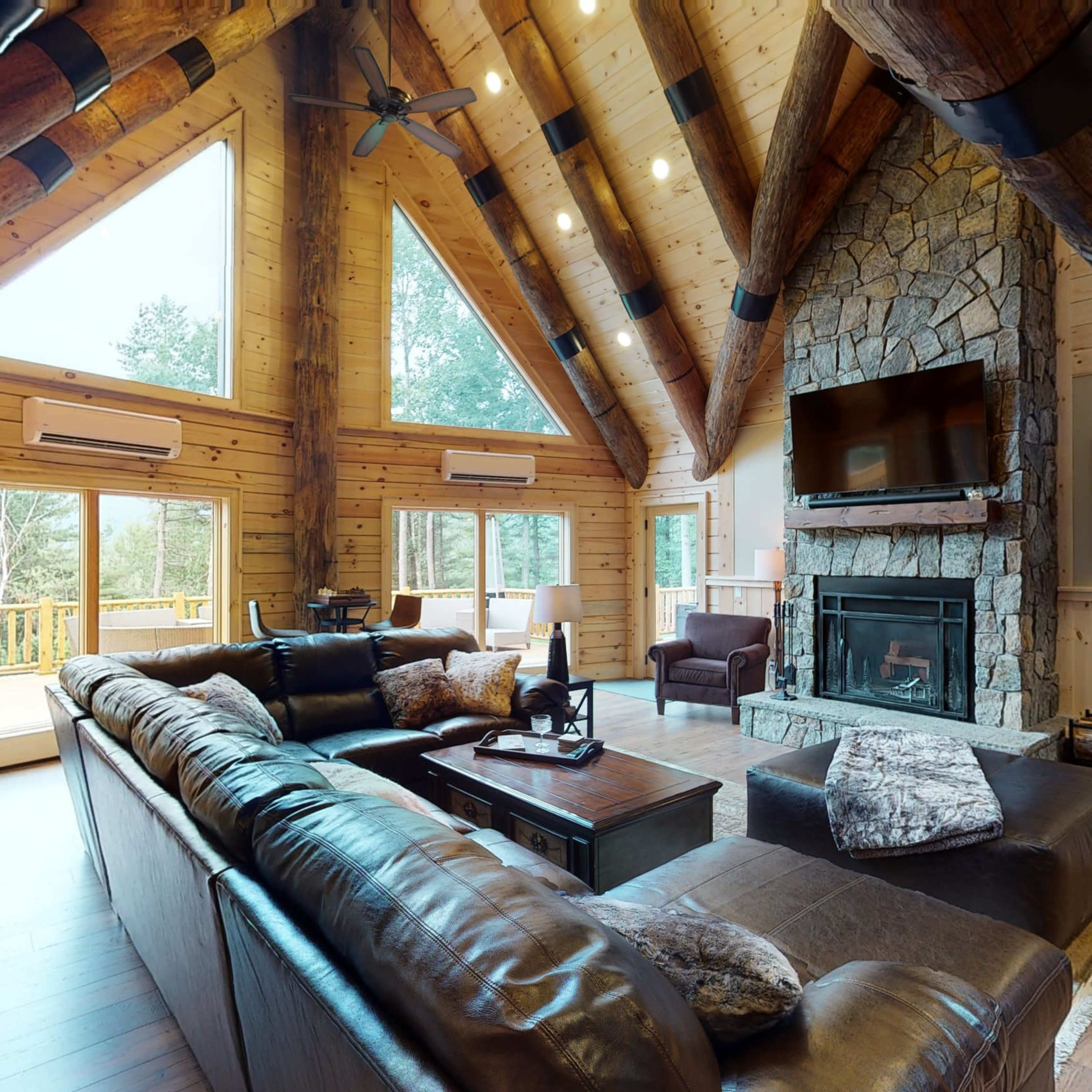 A spacious wooden living room with a stone fireplace, large windows, and vaulted ceilings supported by exposed beams.