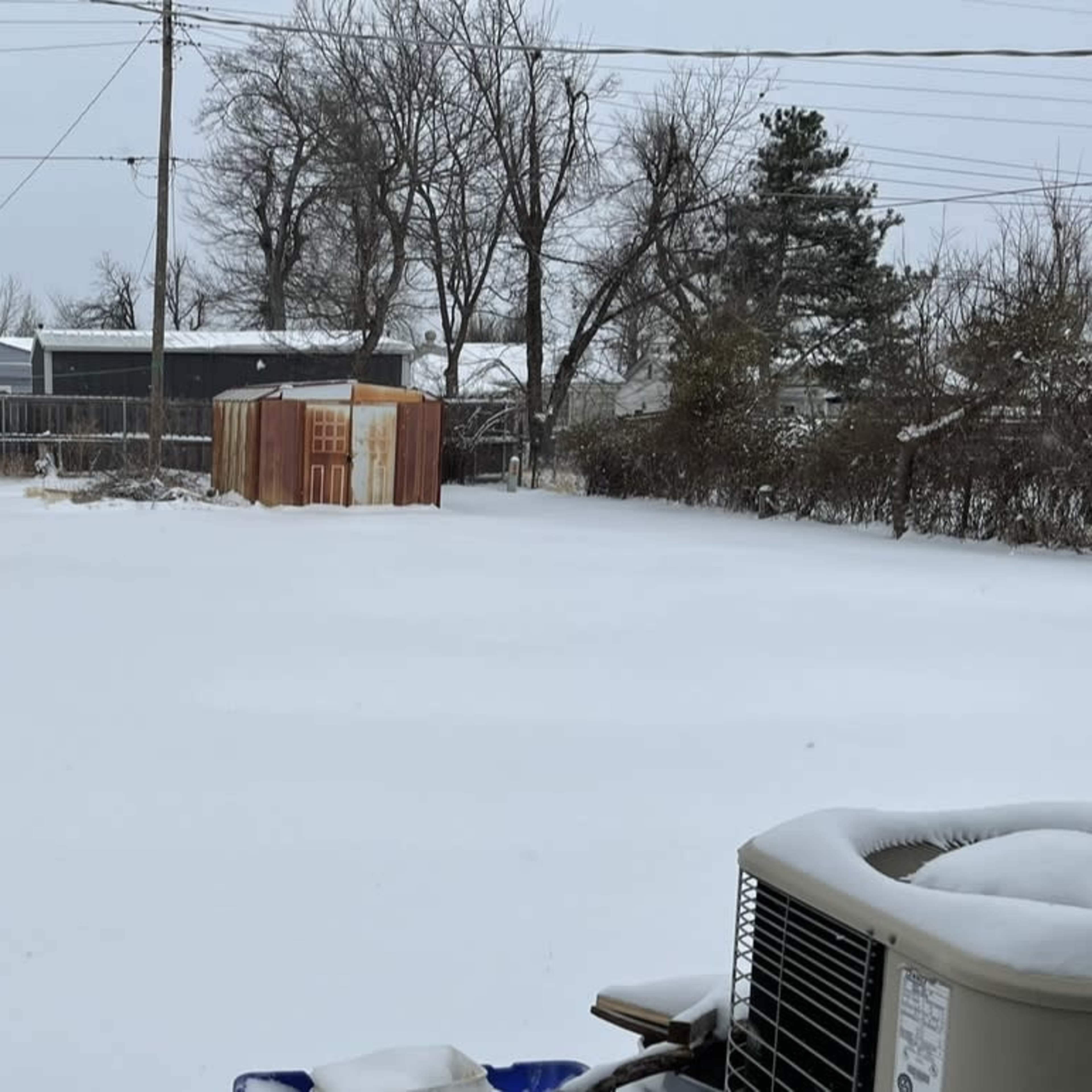 A snow-covered backyard with a shed in the distance and a visible air conditioning unit in the foreground.