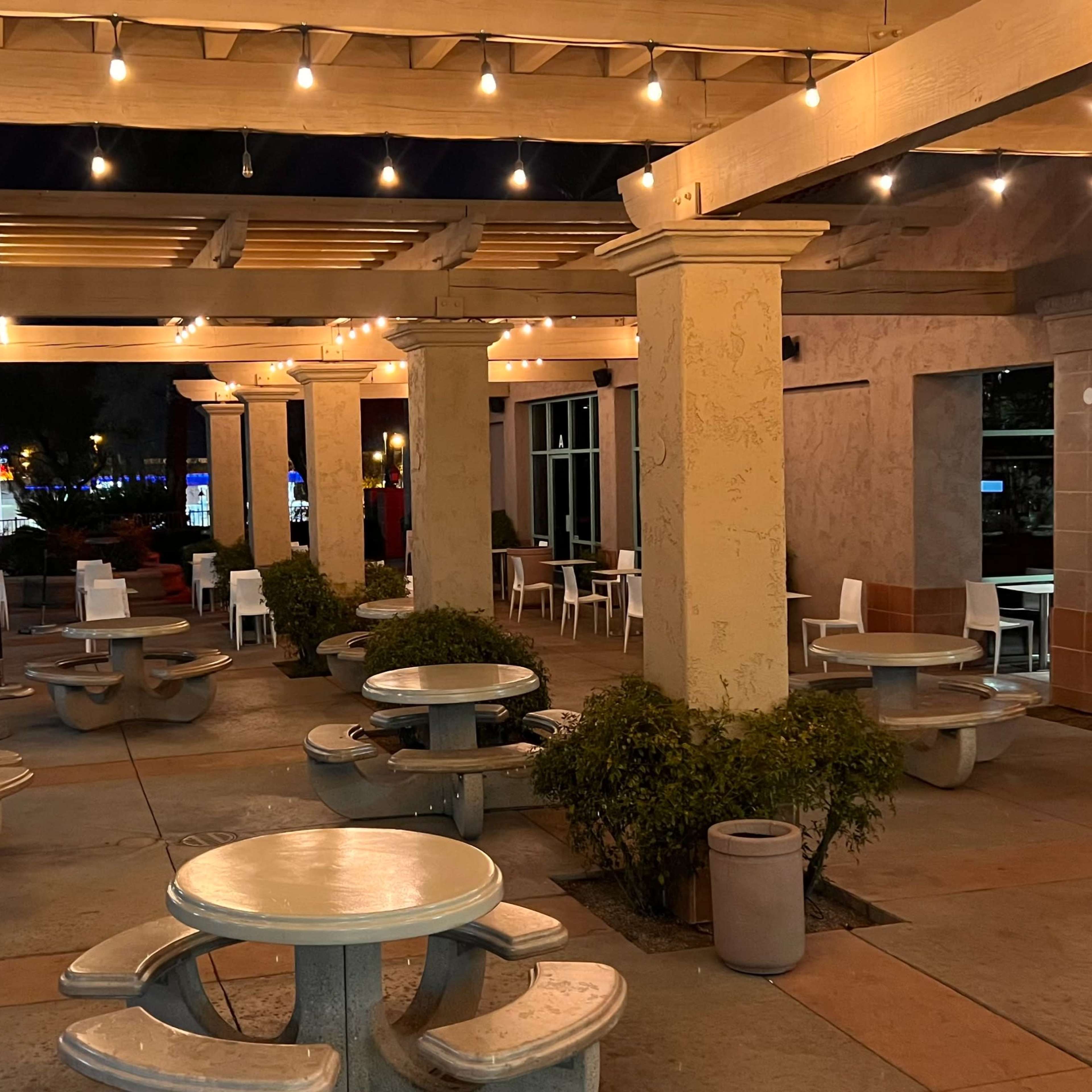 An outdoor dining area at night, complete with circular tables, bench seating, and string lights overhead.