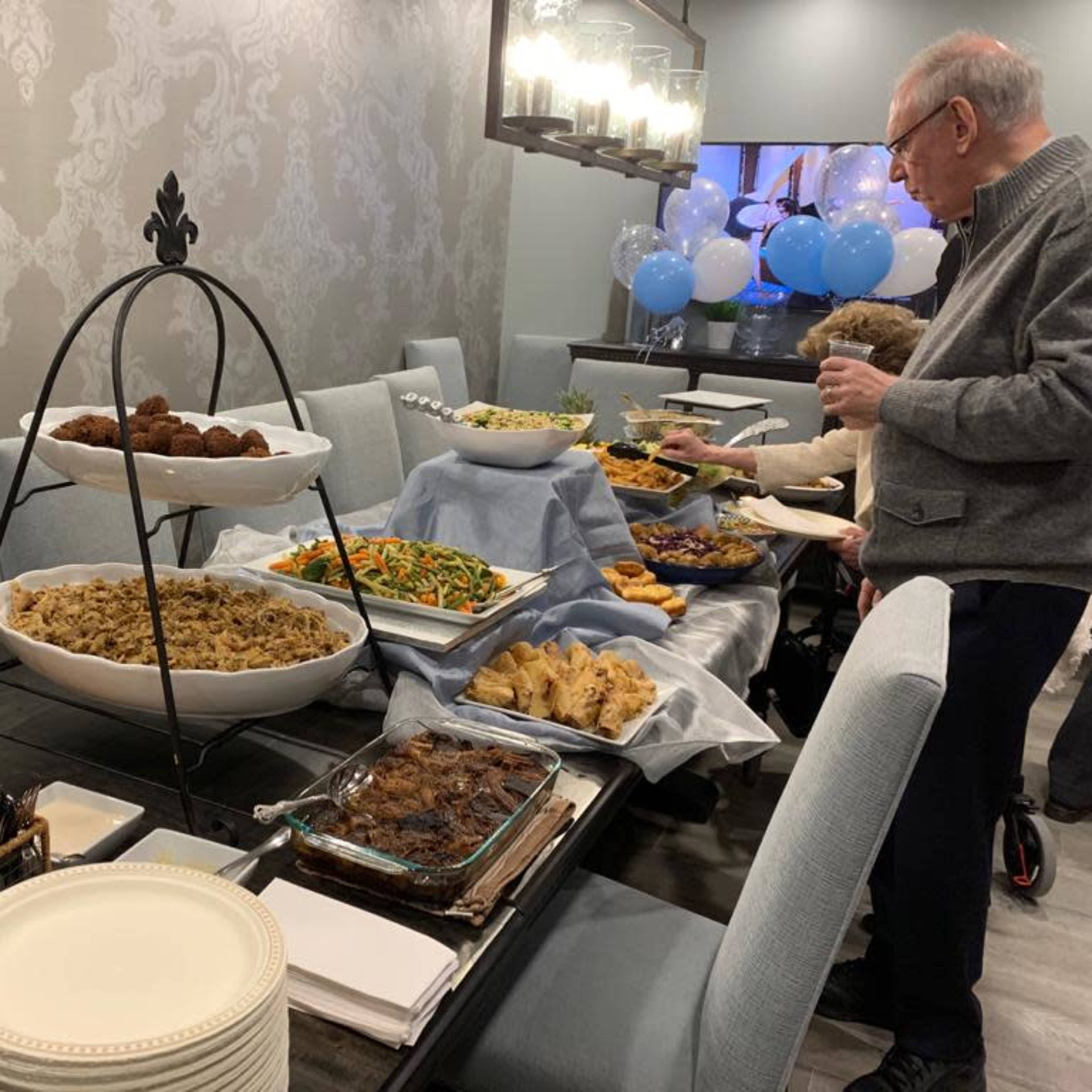 A buffet table is set with various dishes, including bread, salads, and fried foods, while an elderly man stands nearby holding a drink.