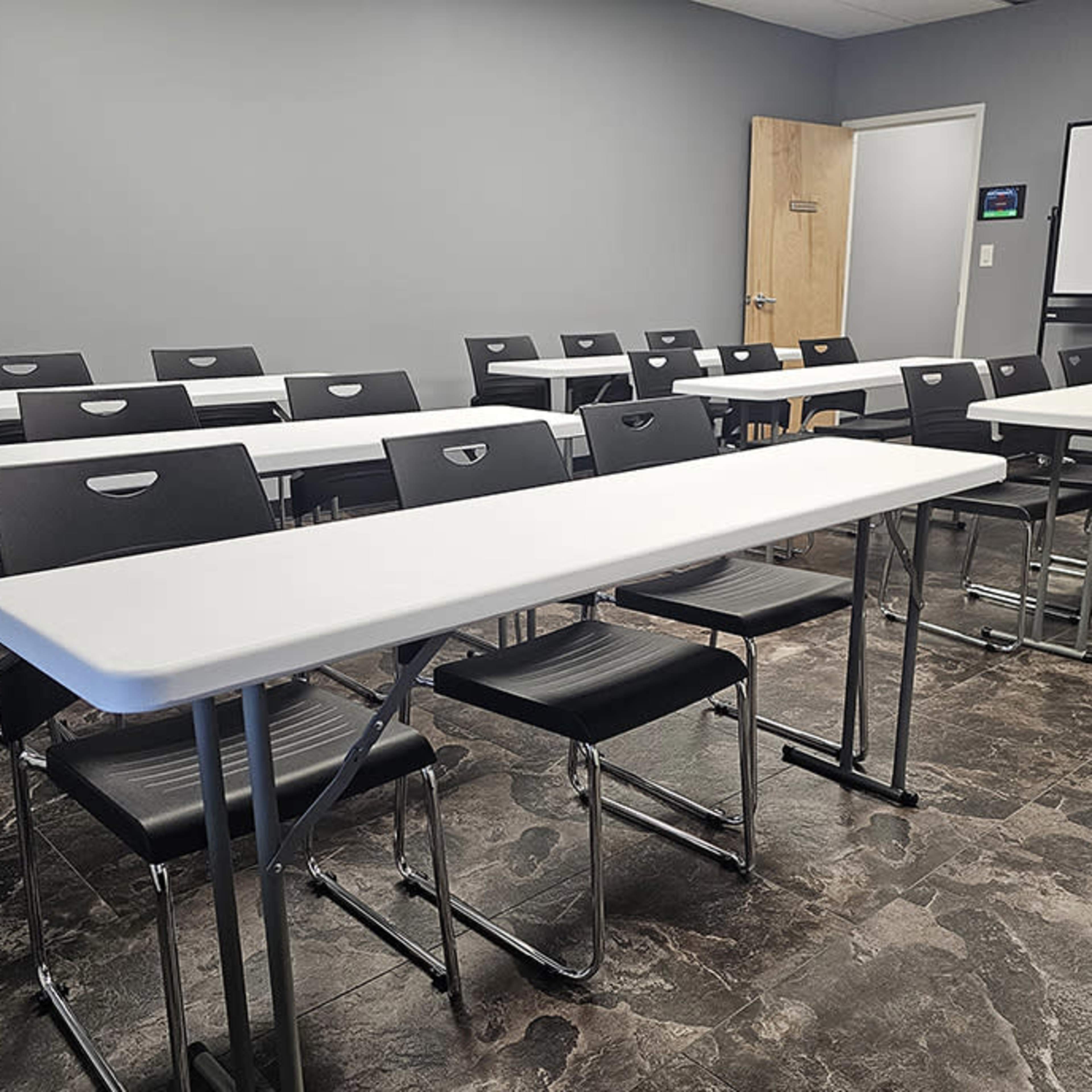 A classroom is set up with several rows of white tables and black chairs arranged neatly against a gray wall, with a screen visible at the front.