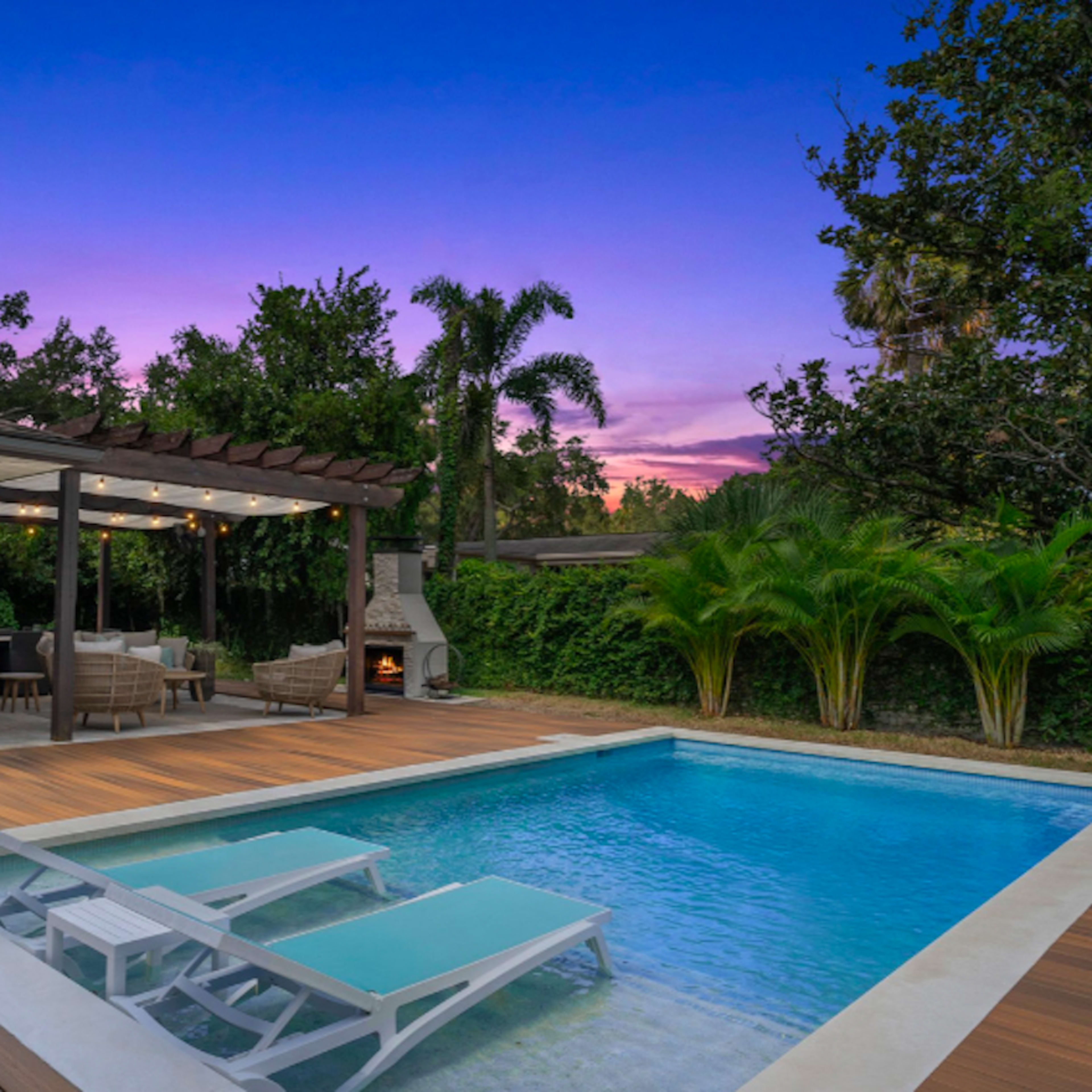 A backyard with a swimming pool, wooden deck, and outdoor seating area under a pergola, set against a sunset sky.