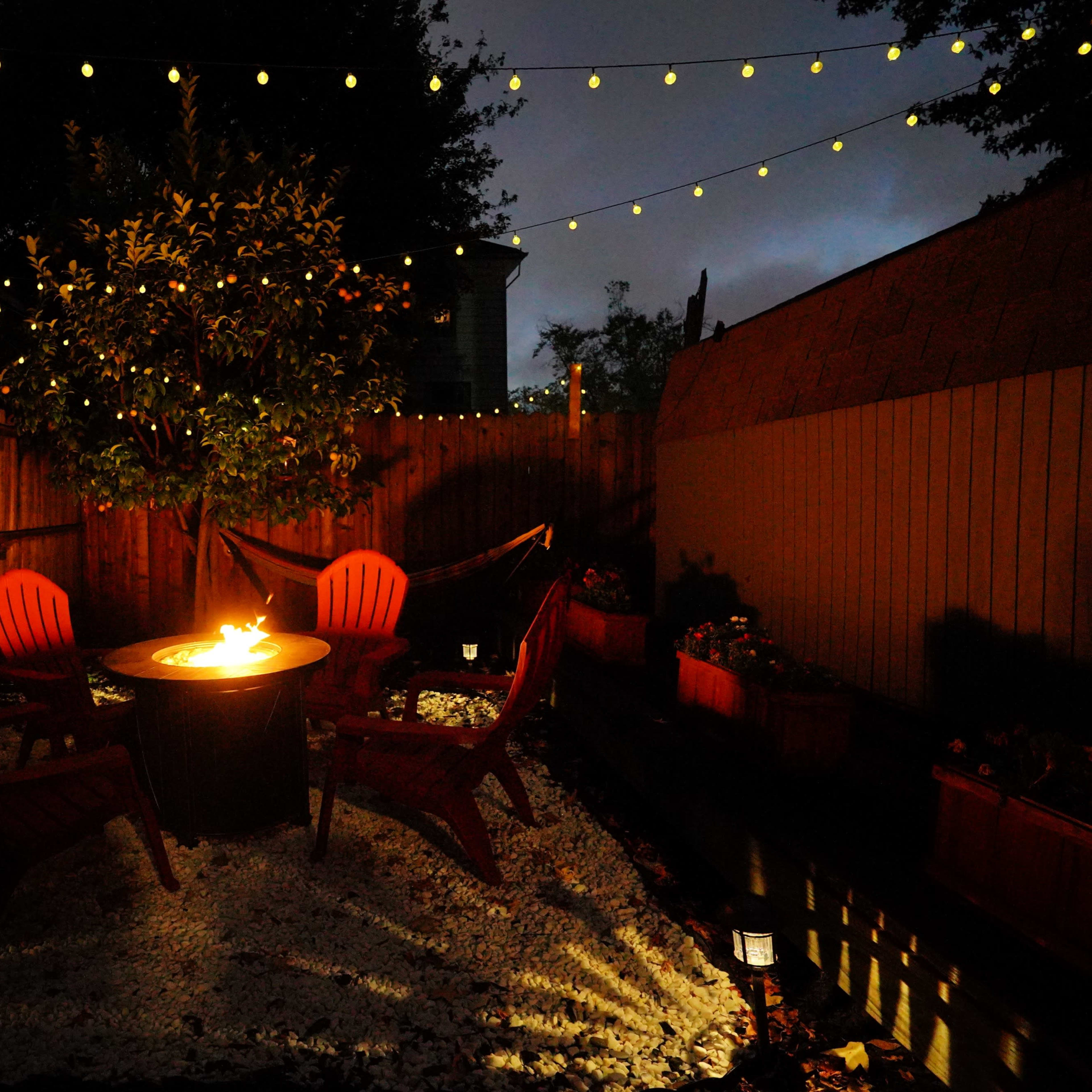 A circular fire pit surrounded by four red chairs is illuminated under string lights in a backyard garden at night.