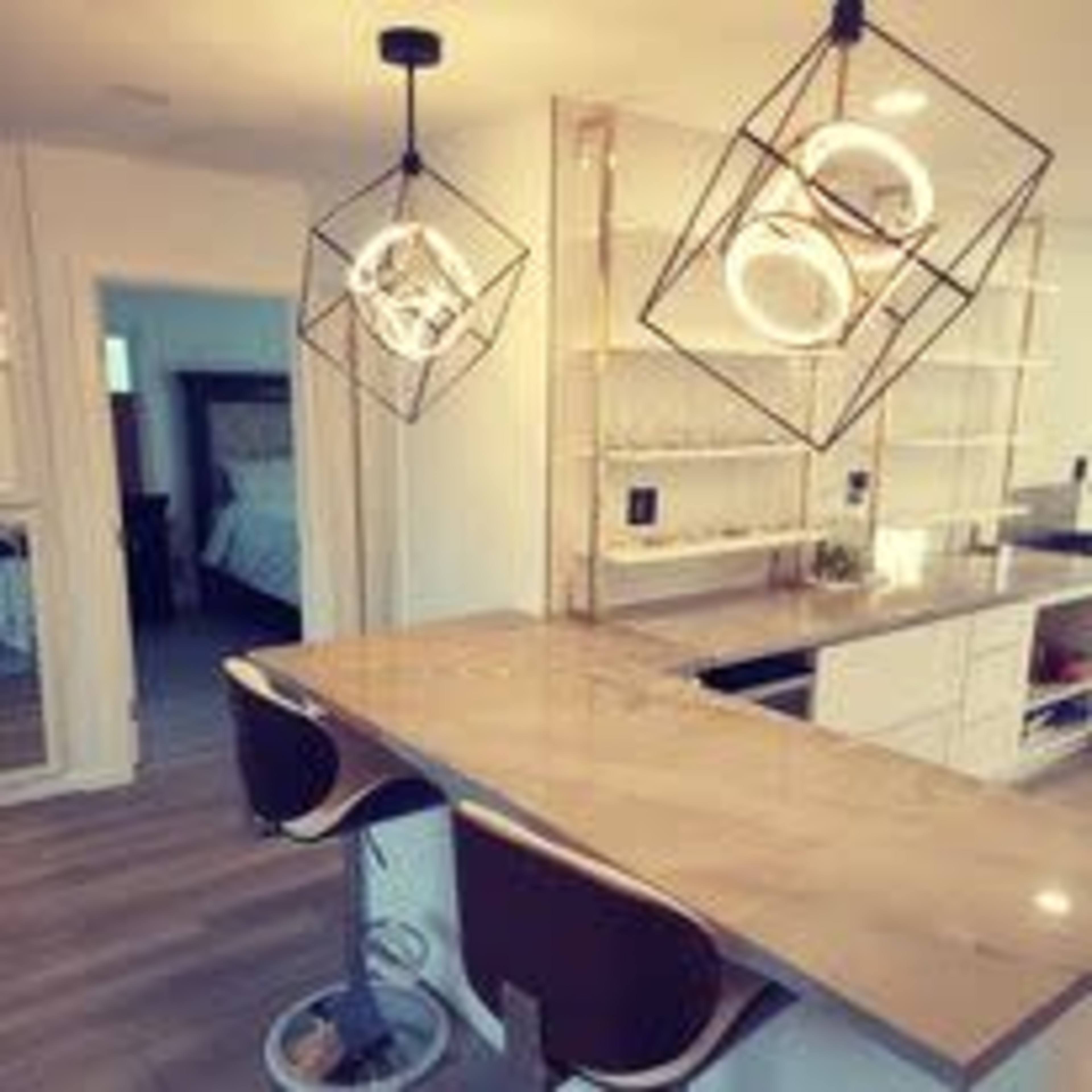 A modern kitchen with two geometric pendant lights hanging above a marble-topped island and stylish bar stools, with an open view to a bedroom in the background.