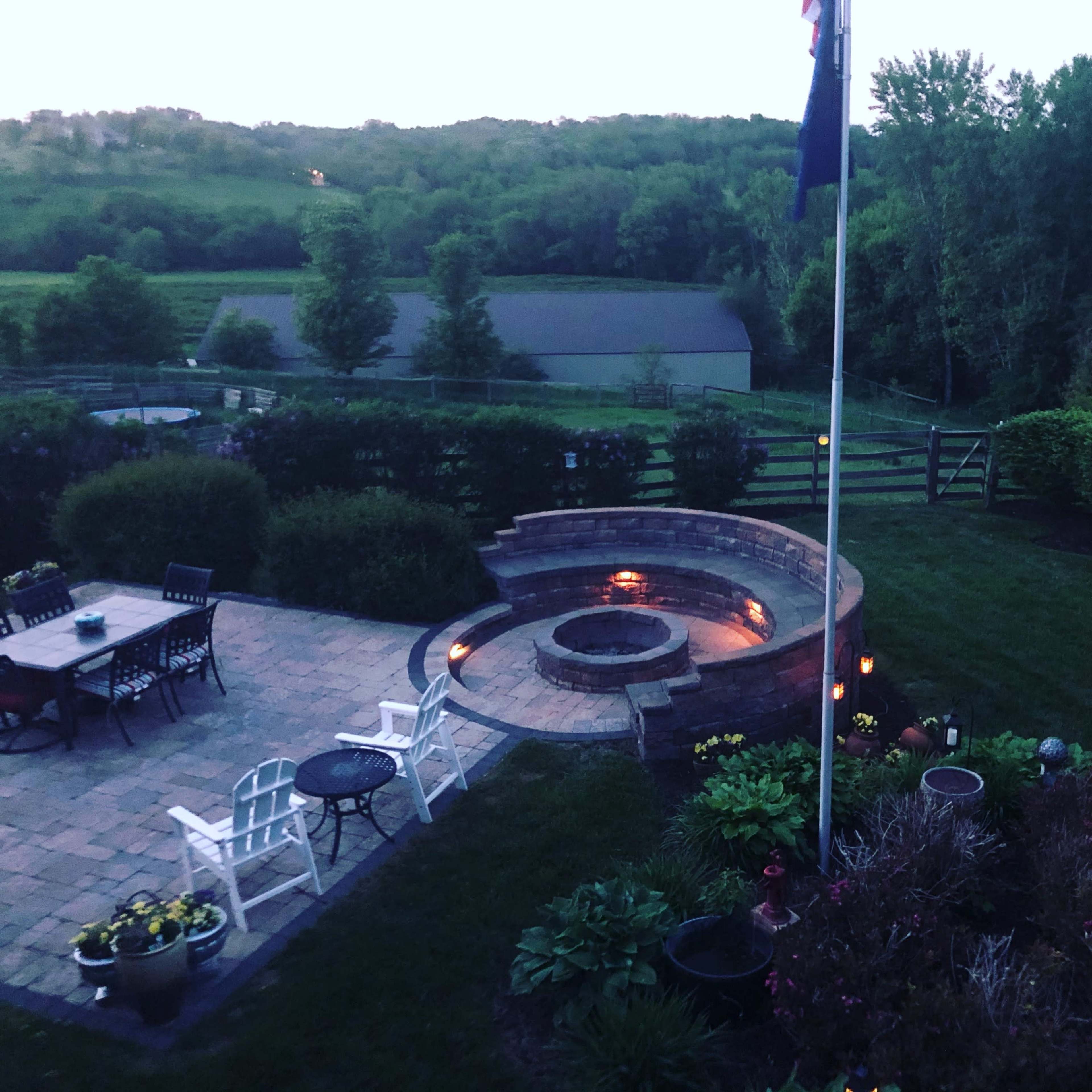 A backyard with a stone fire pit surrounded by seating, a dining area with chairs and a table, and landscaped greenery under evening light.