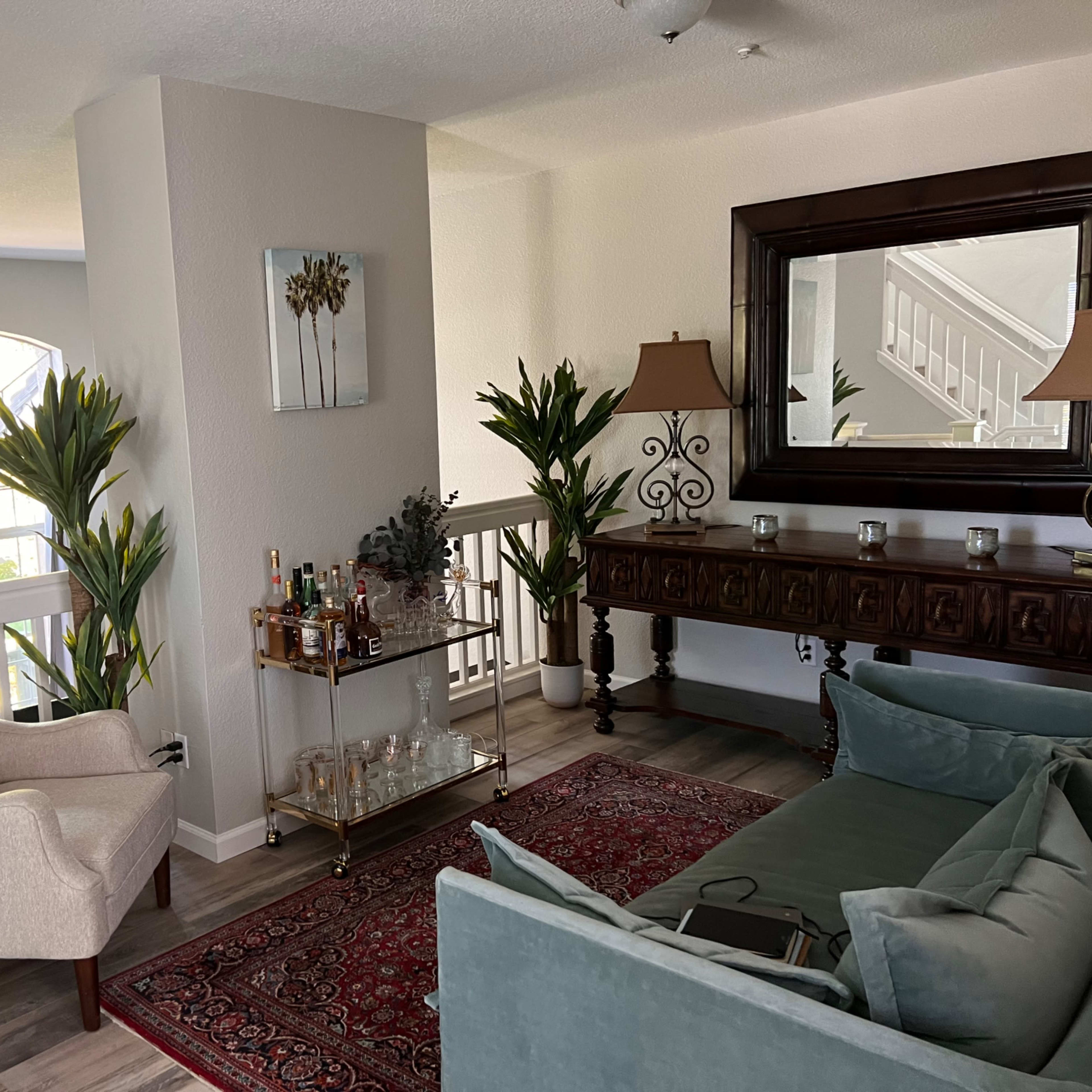 A living room with a sofa, an armchair, a wooden console table with decorative items, and tropical plants, arranged around a small area rug.