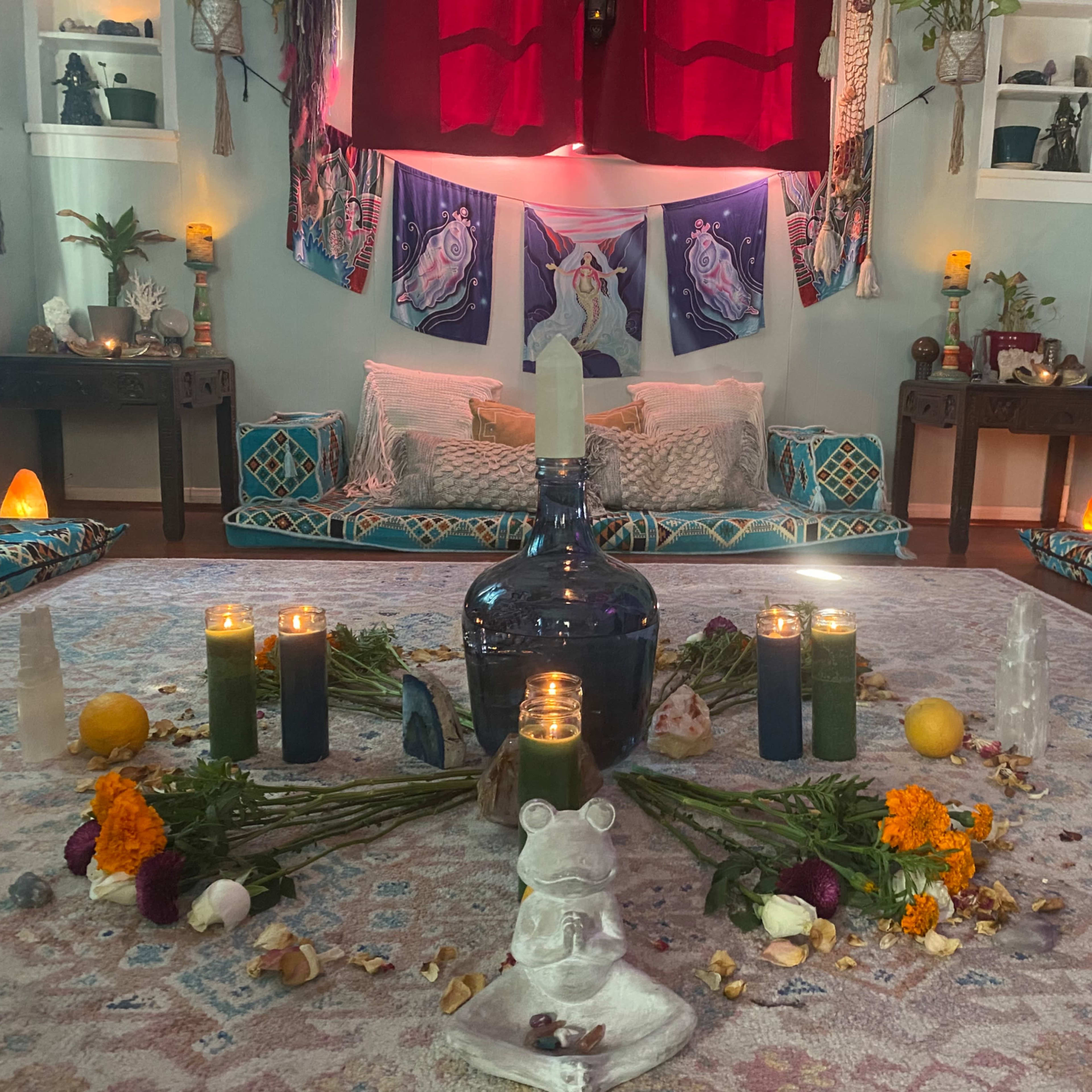 A cozy, well-decorated yoga or meditation space with cushions, candles, flowers, and an ornate centerpiece on a patterned rug.