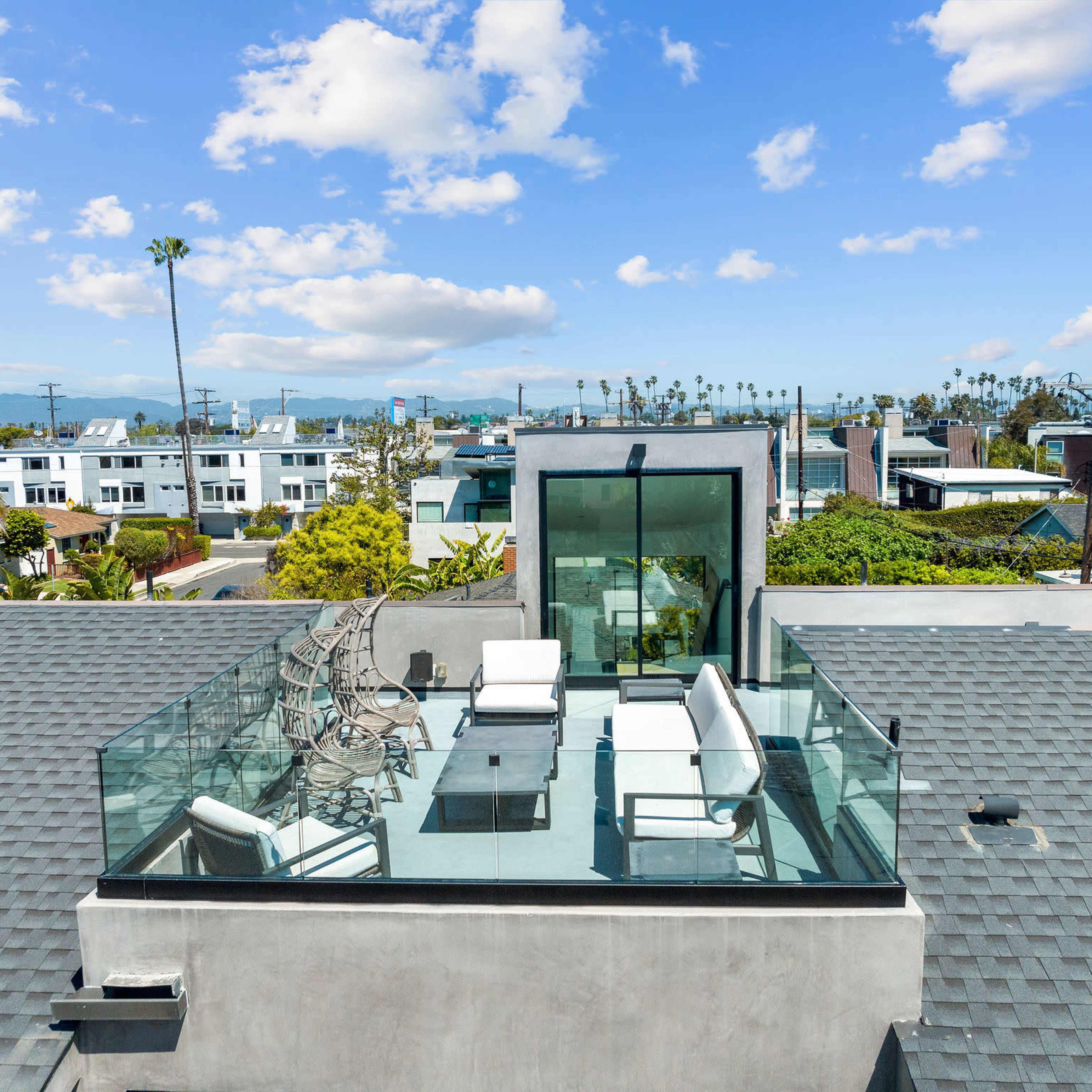 A rooftop terrace features white outdoor furniture and a glass railing, overlooking a coastal cityscape with palm trees and distant mountains.