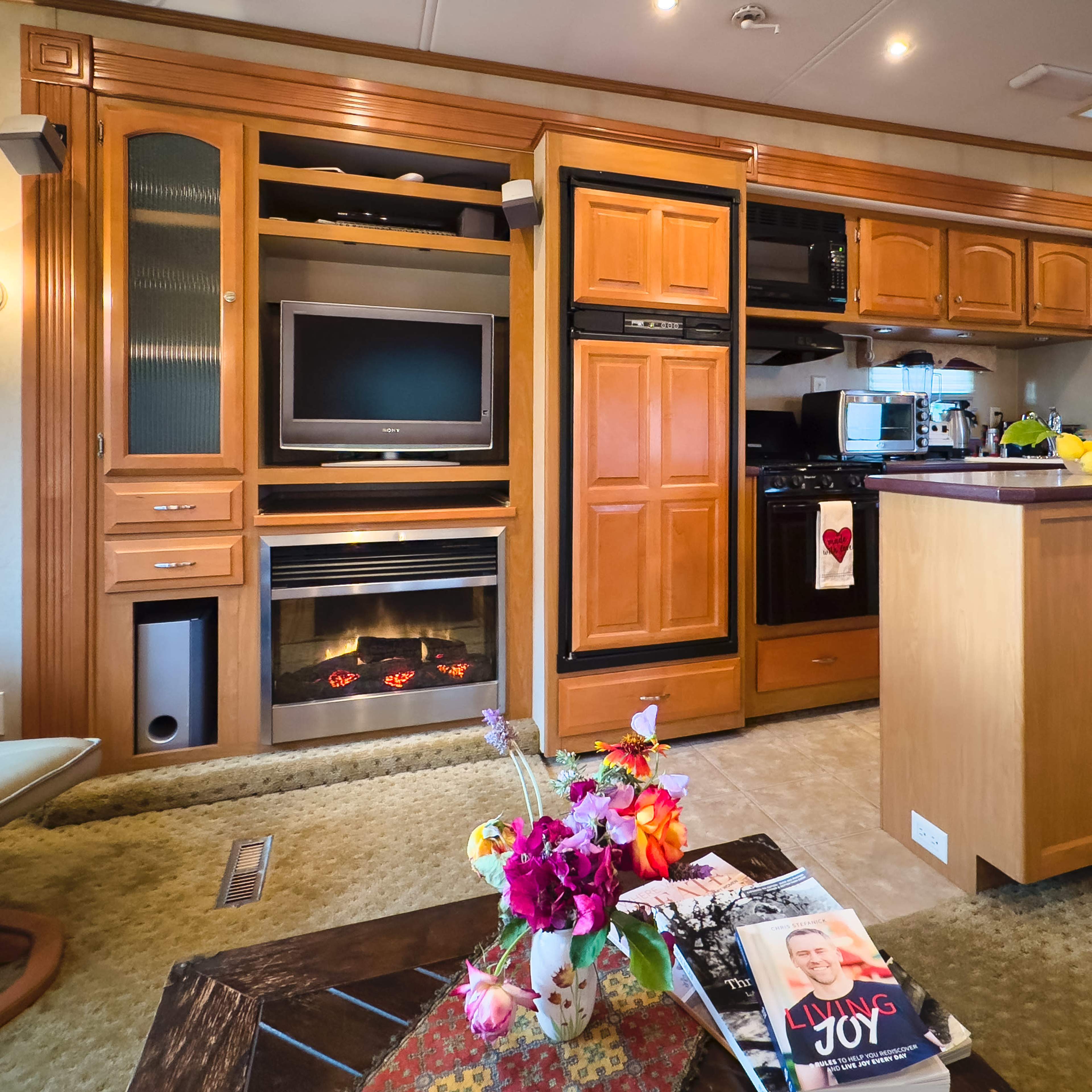 A cozy kitchen and living area with wooden cabinetry, a television, and a kitchen island, decorated with a vase of flowers and magazines on a coffee table.