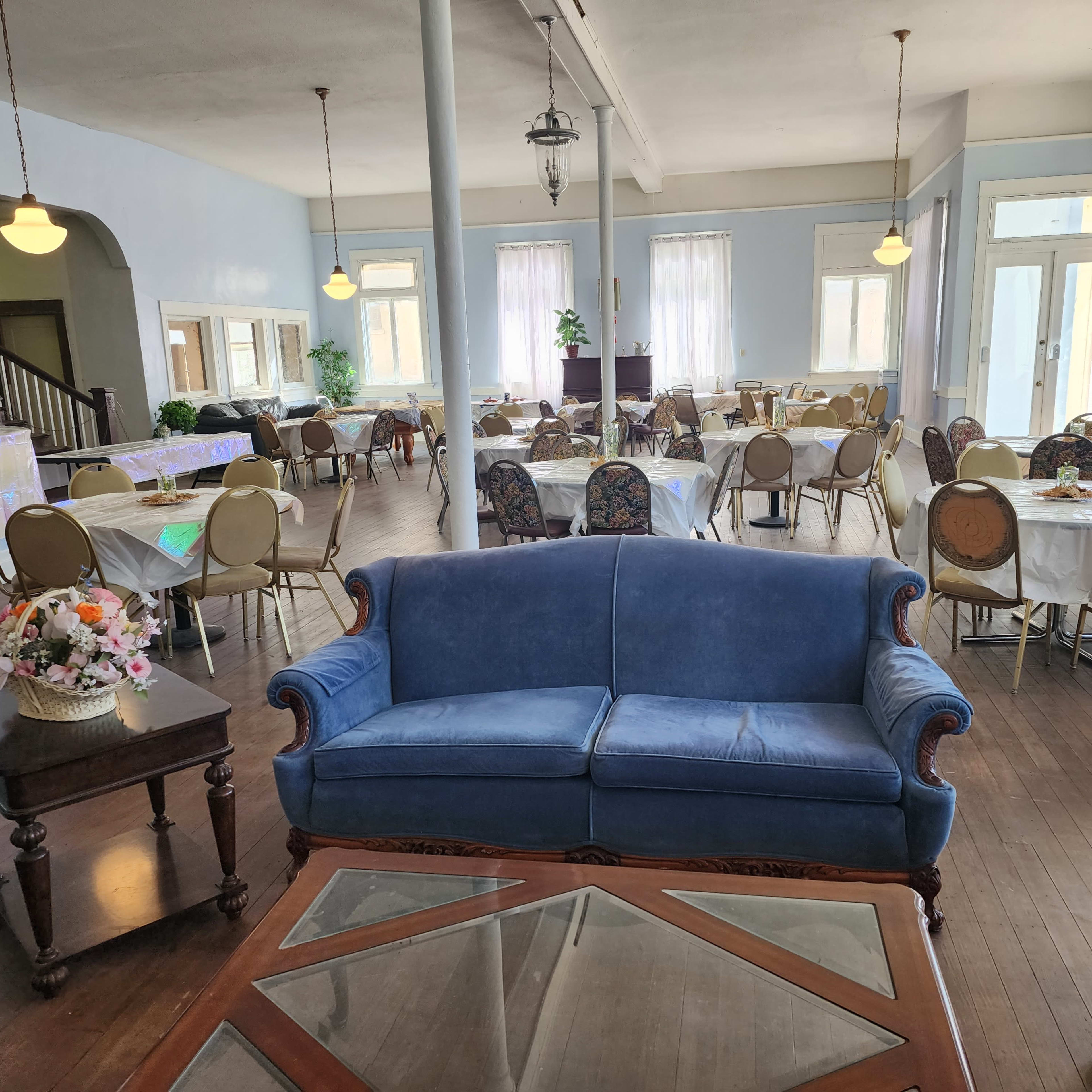 A spacious dining area with tables set for meals, a blue sofa in the foreground, and large windows allowing natural light to enter.
