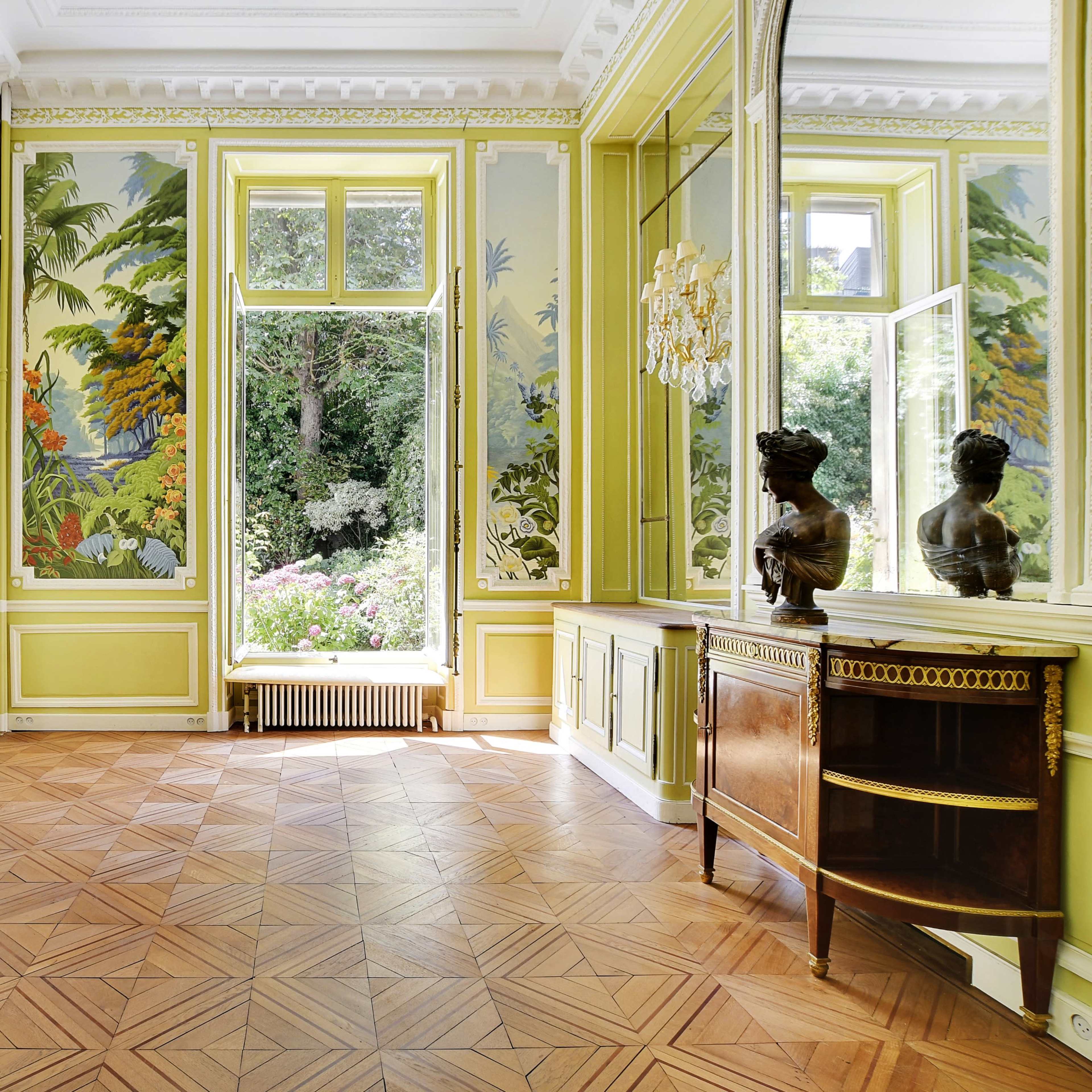 The room features elegant yellow walls adorned with botanical murals, large windows overlooking a garden, and wooden furniture arranged along the walls.