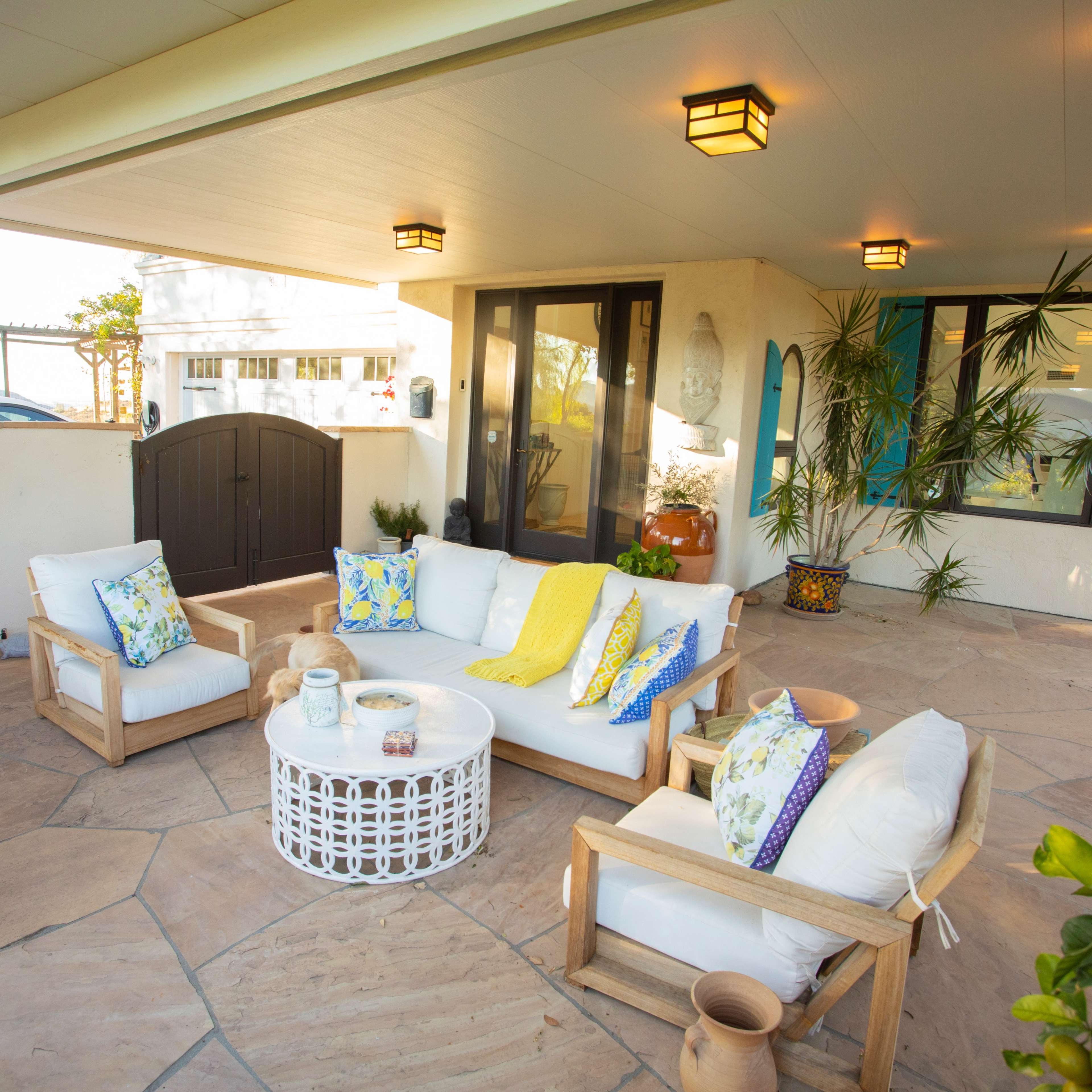 A cozy outdoor seating area with a white couch and wooden chairs, arranged around a round coffee table on a stone patio.