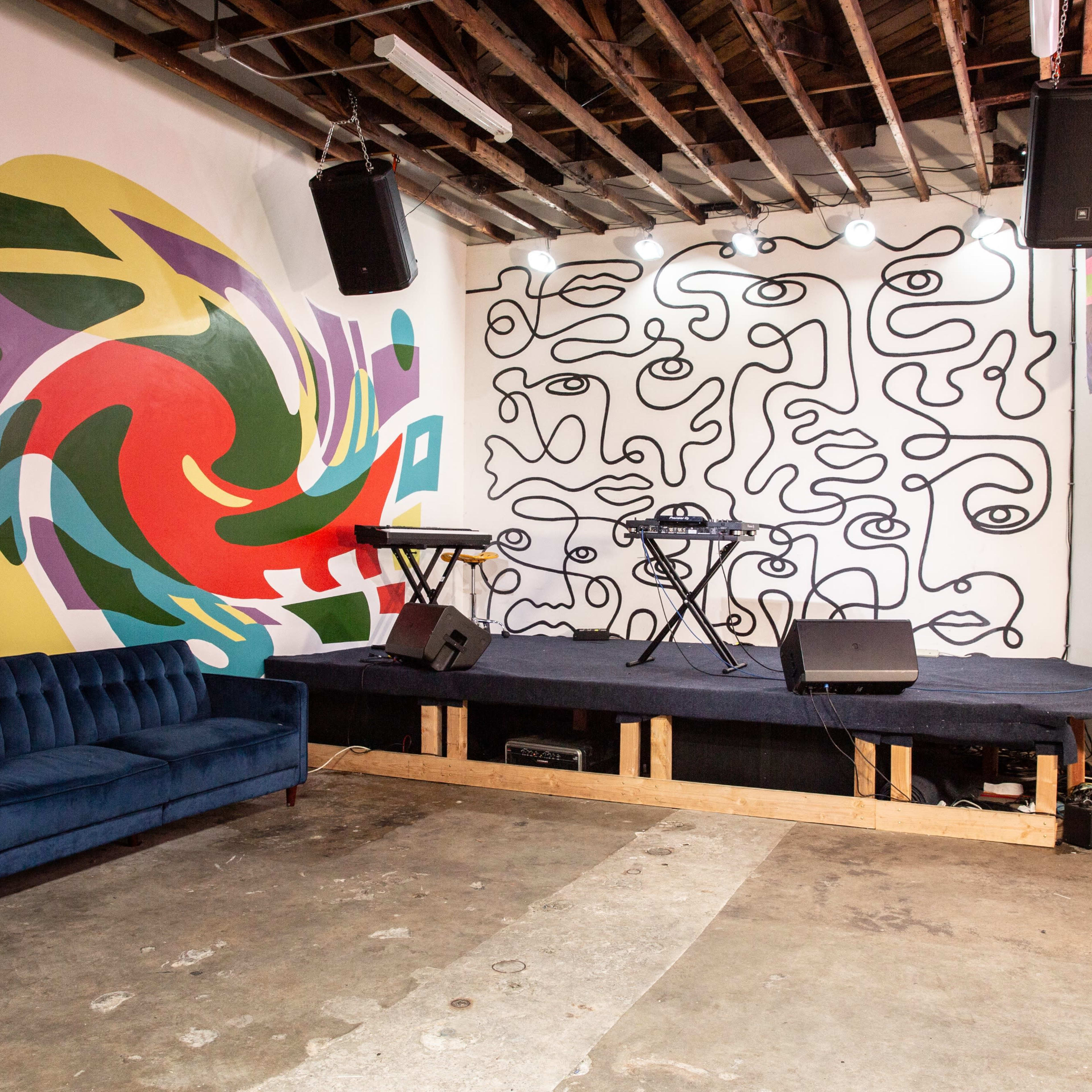 A small performance space featuring a stage with two keyboards, speakers mounted on the wall, and colorful murals on the adjacent walls.