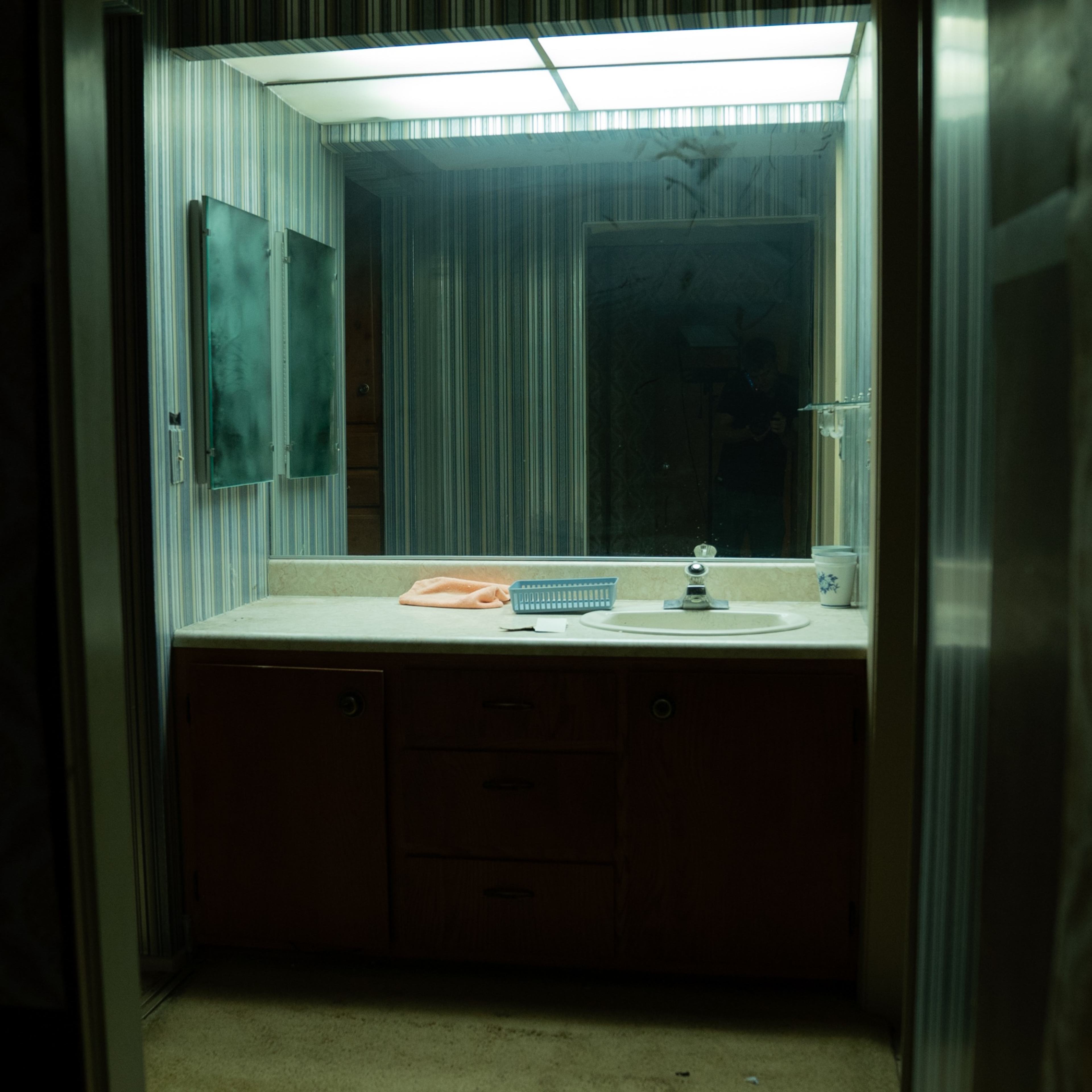 A dimly lit bathroom with a sink, a countertop, and a large mirror surrounded by striped wallpaper.