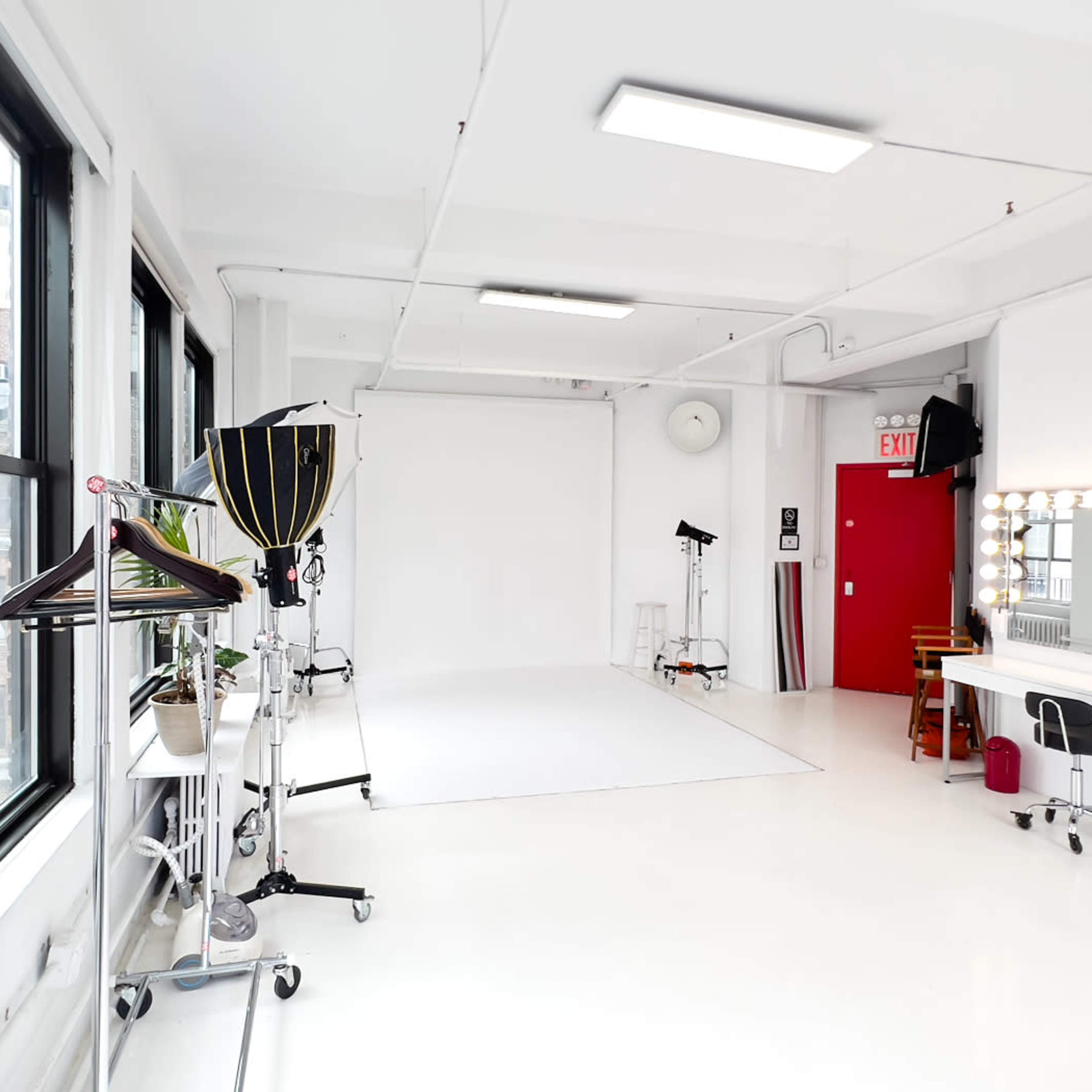 A bright, minimally furnished photography studio featuring a white backdrop, dressing area with mirrors, and a few equipment stands.