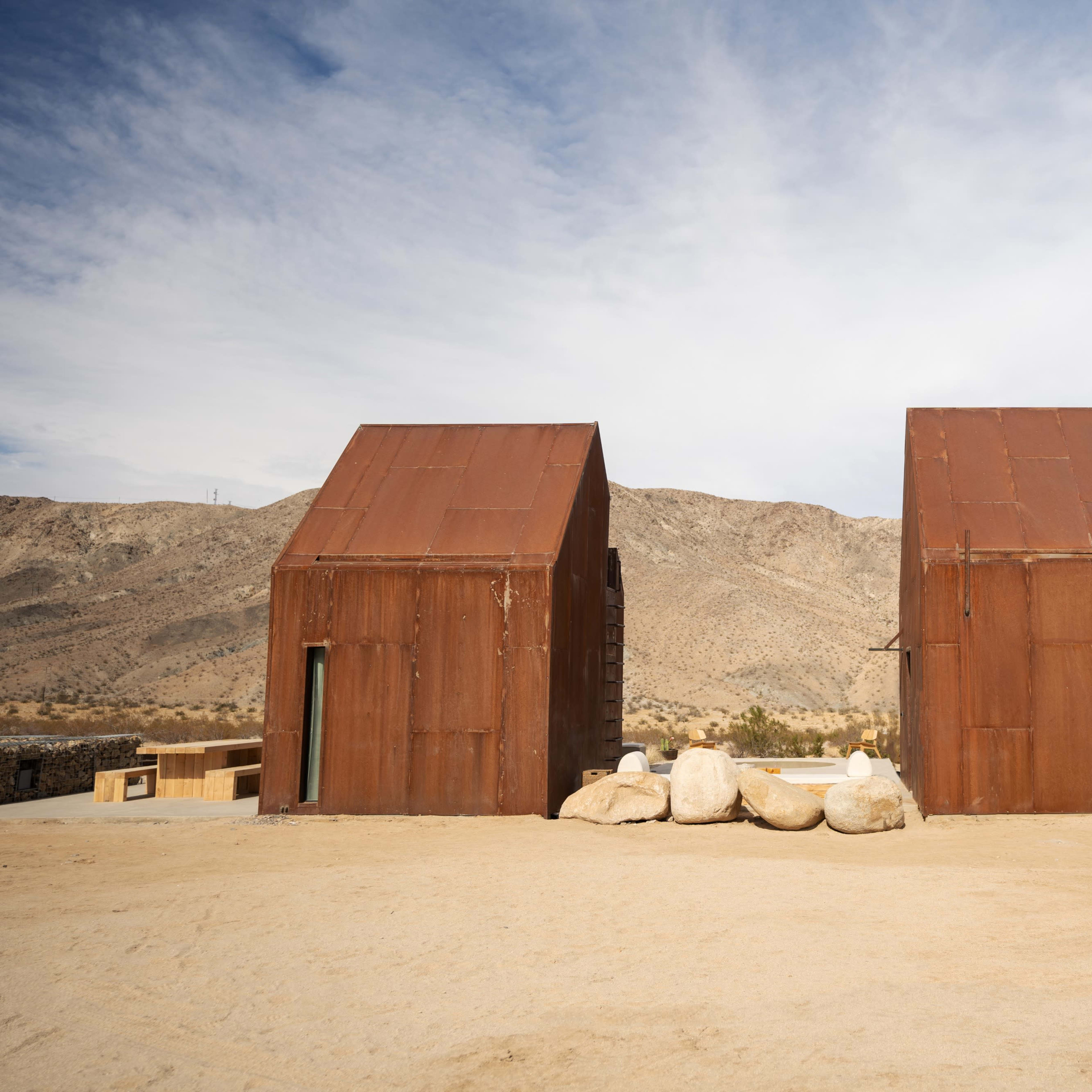 Two rust-colored metal houses are situated in a desert landscape, with rocky terrain and a mountain backdrop.