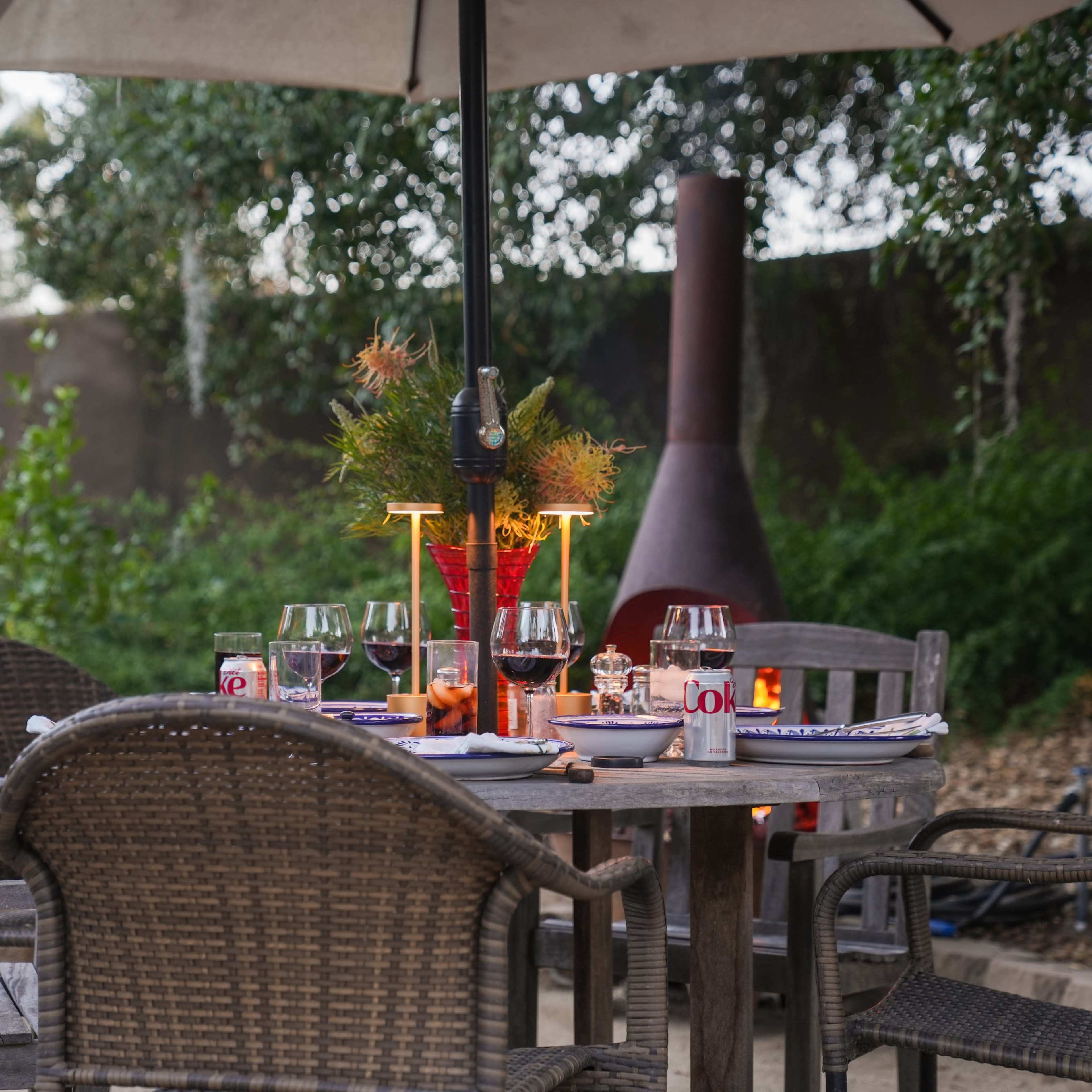 A dining table is set outdoors under a large umbrella, surrounded by chairs and adorned with glasses, candles, and decorative elements.